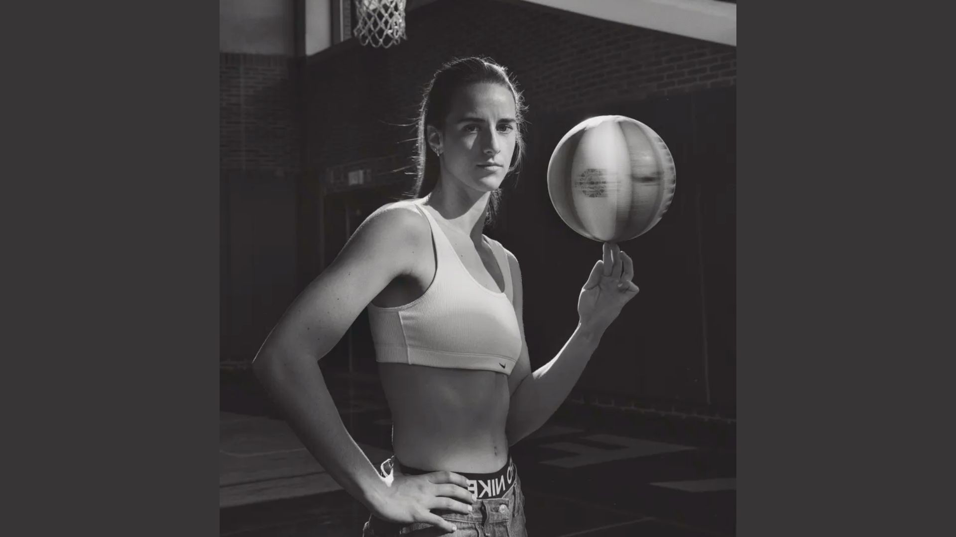 "Black and white image of a confident woman in a sports bra, spinning a basketball on her finger on an indoor court. She stands with poise and determination