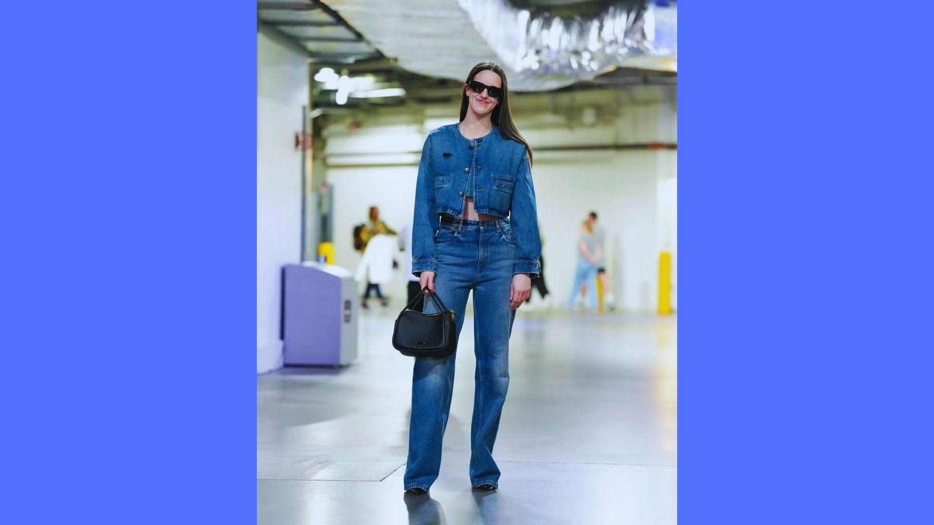 A woman in a denim outfit and sunglasses walks confidently through a modern, well-lit hallway. She carries a black handbag, exuding a stylish, casual vibe.