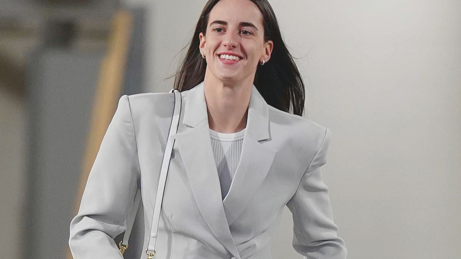 "A person in a light gray tailored suit walks confidently indoors, smiling. They carry a white bag, exuding a professional and cheerful vibe.