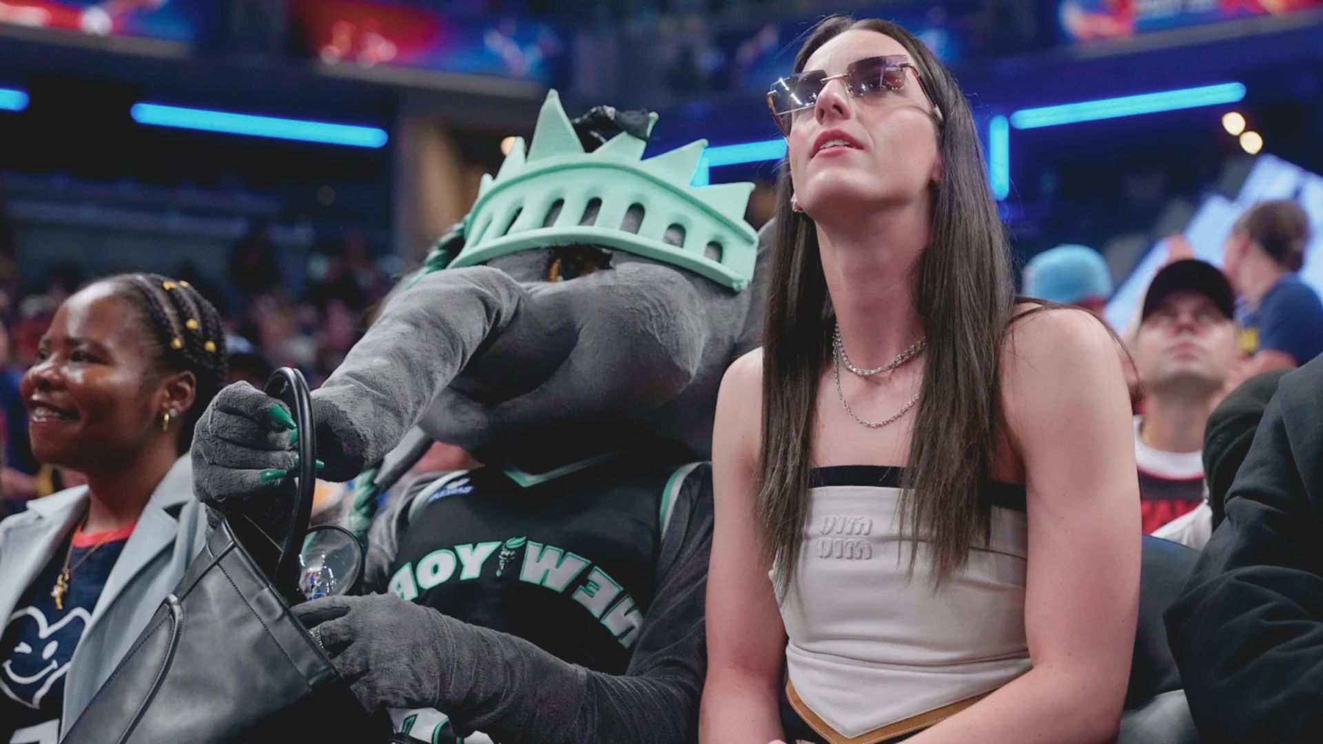 A woman in sunglasses sits courtside at a lively basketball game next to a person in a playful mascot costume, creating a lively, fun atmosphere.