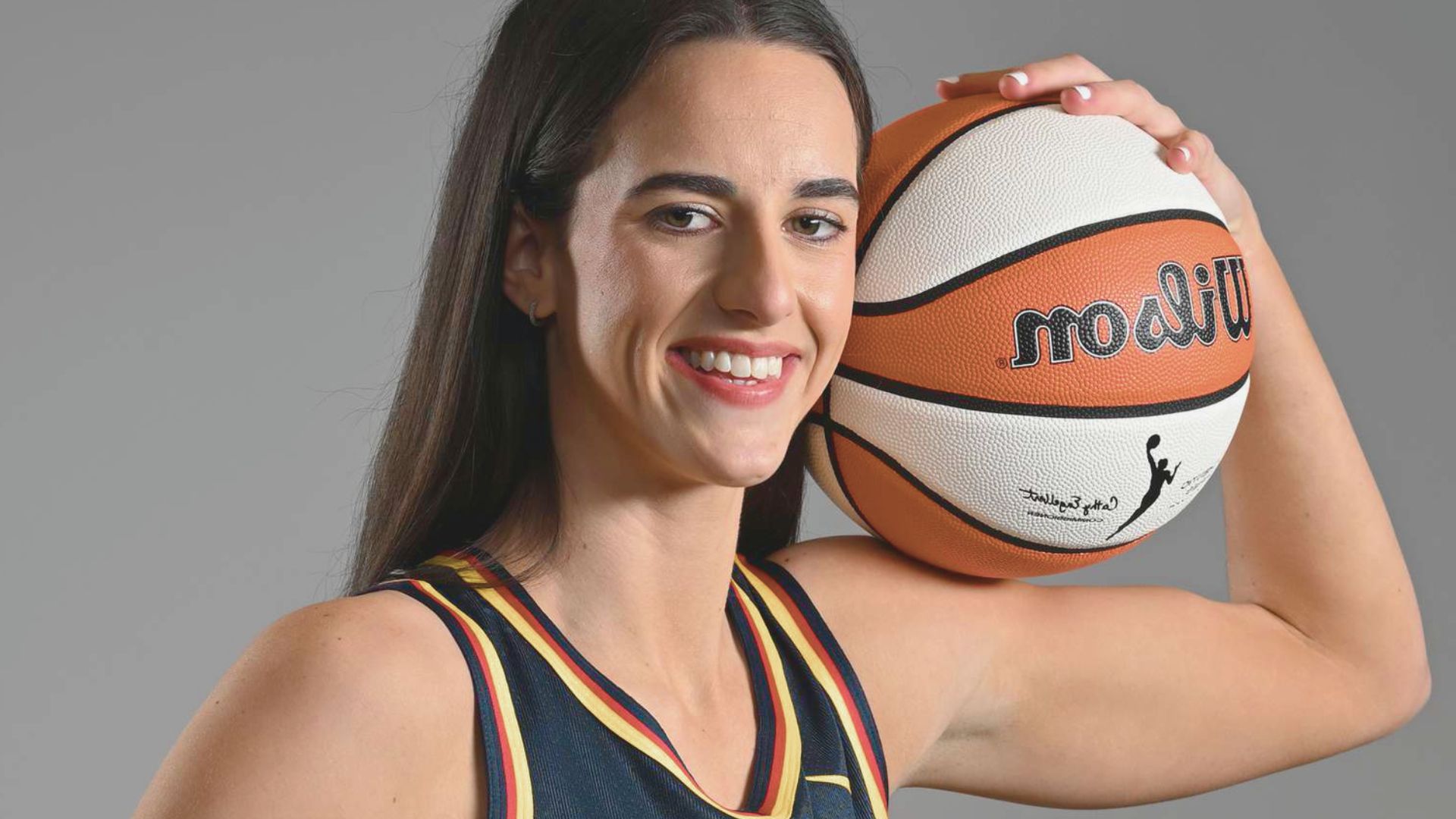 Woman in a basketball jersey smiles confidently, resting a basketball on her shoulder. The background is gray, emphasizing her sporty and cheerful demeanor.
