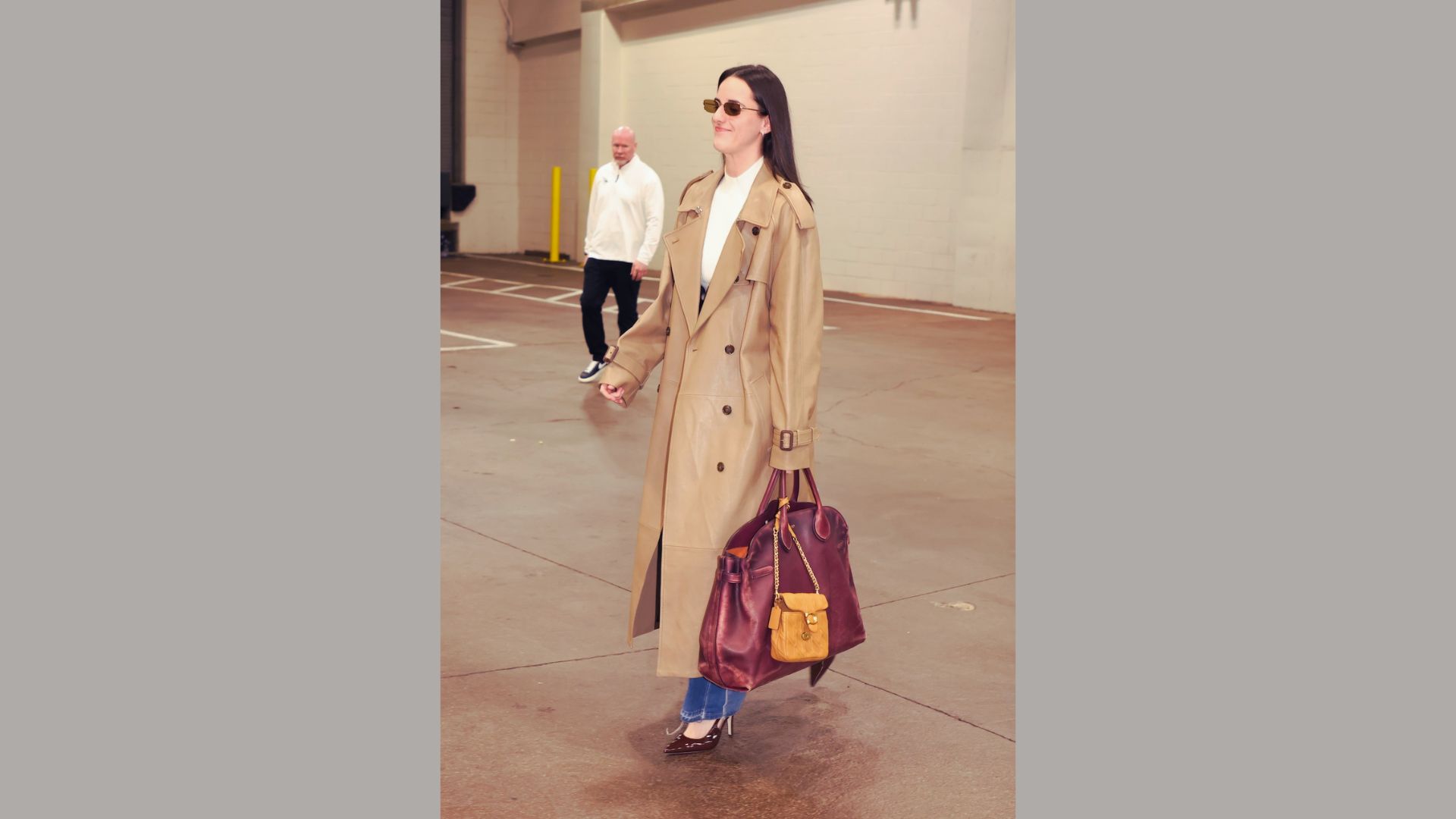 "A woman in a beige trench coat and sunglasses walks confidently on a street, holding a large burgundy handbag. The background features an urban setting.