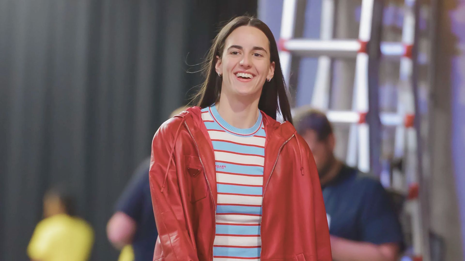 A person in a red jacket and striped shirt smiles warmly. They are in front of a blurred background with a ladder and figures, creating a lively atmosphere.