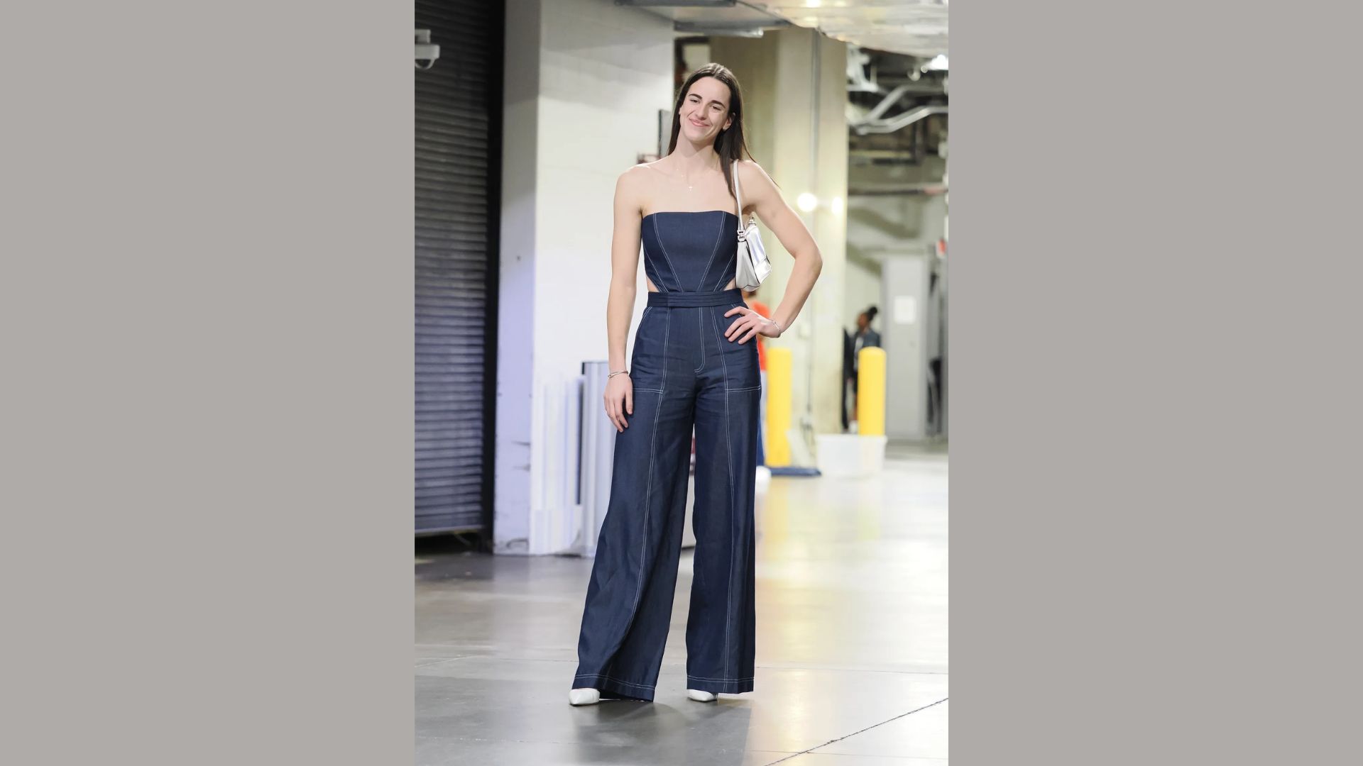 A woman in a strapless denim jumpsuit stands confidently in an industrial hallway, with one hand on her hip. The space is lit brightly, evoking a casual yet stylish vibe.