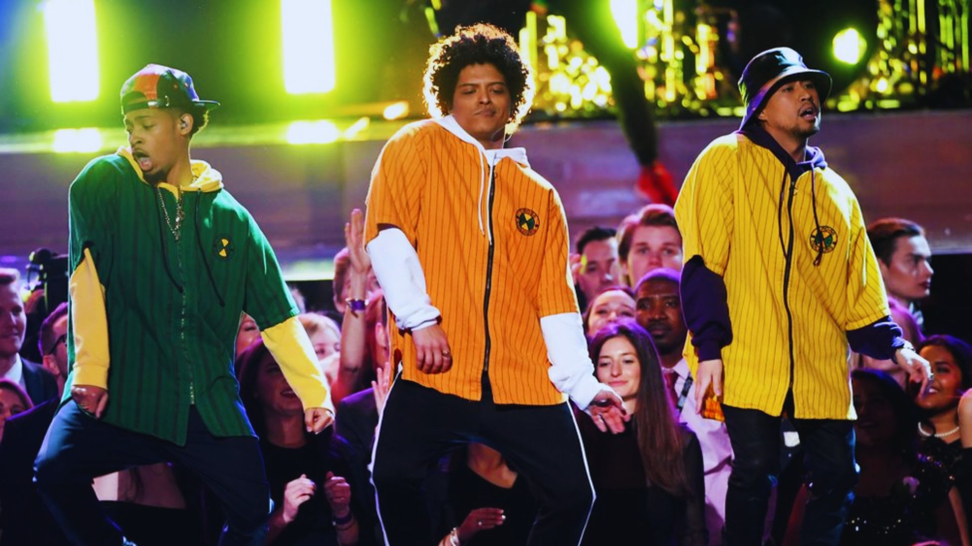 Three people on stage, including Bruno Mars in an orange hoodie, probably performing in a concert.