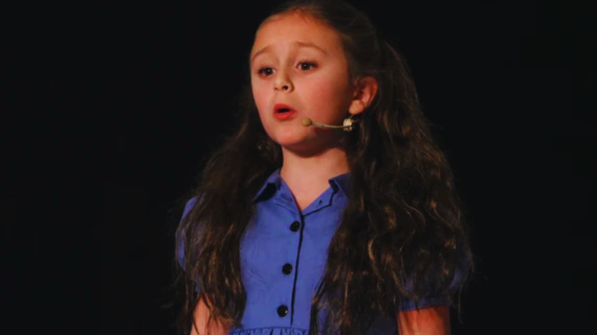 A young girl with long hair singing into a microphone, expressing joy and enthusiasm.