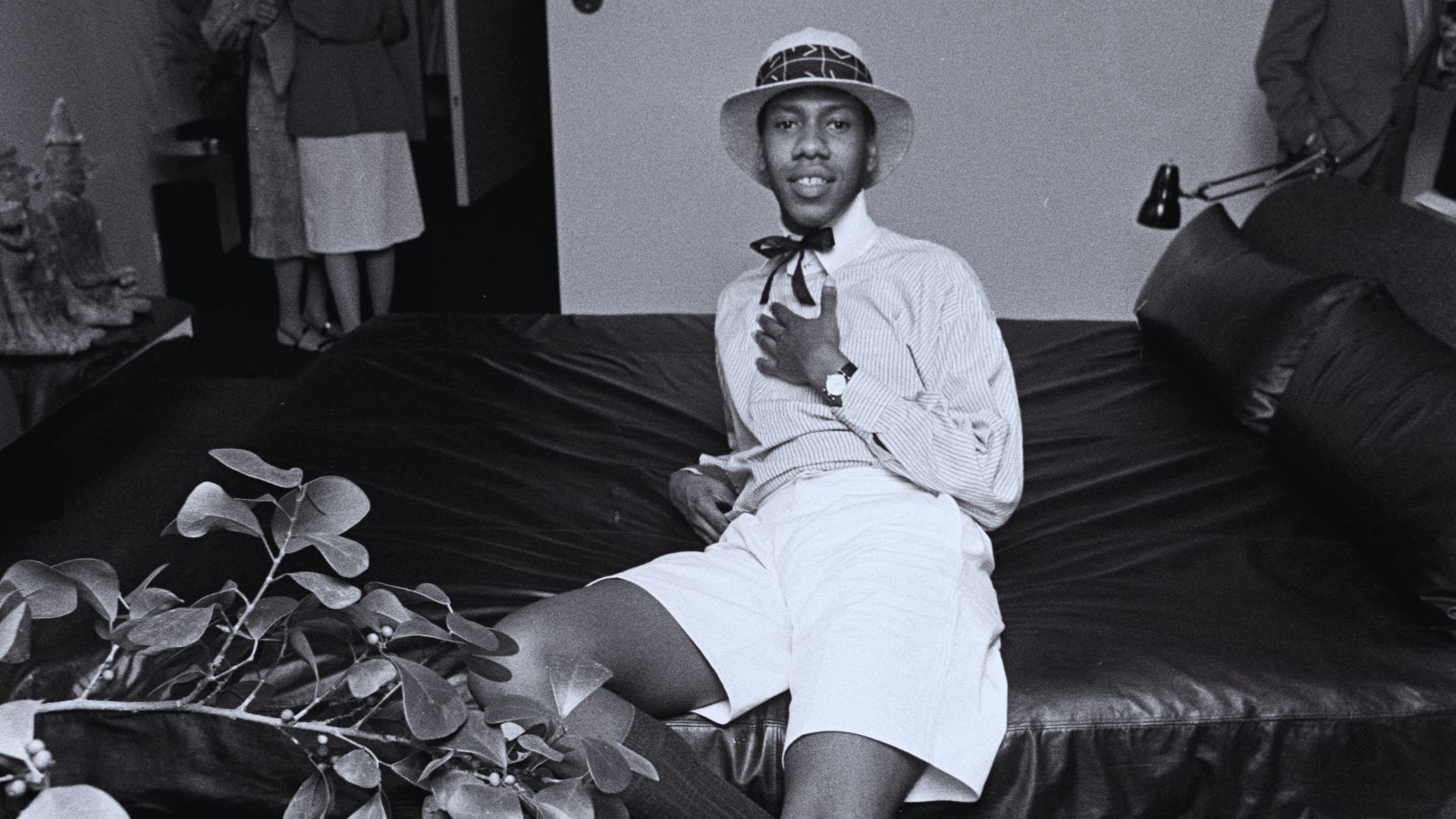 A man in a white shirt and tie sits on a bed, looking contemplative in a softly lit room.