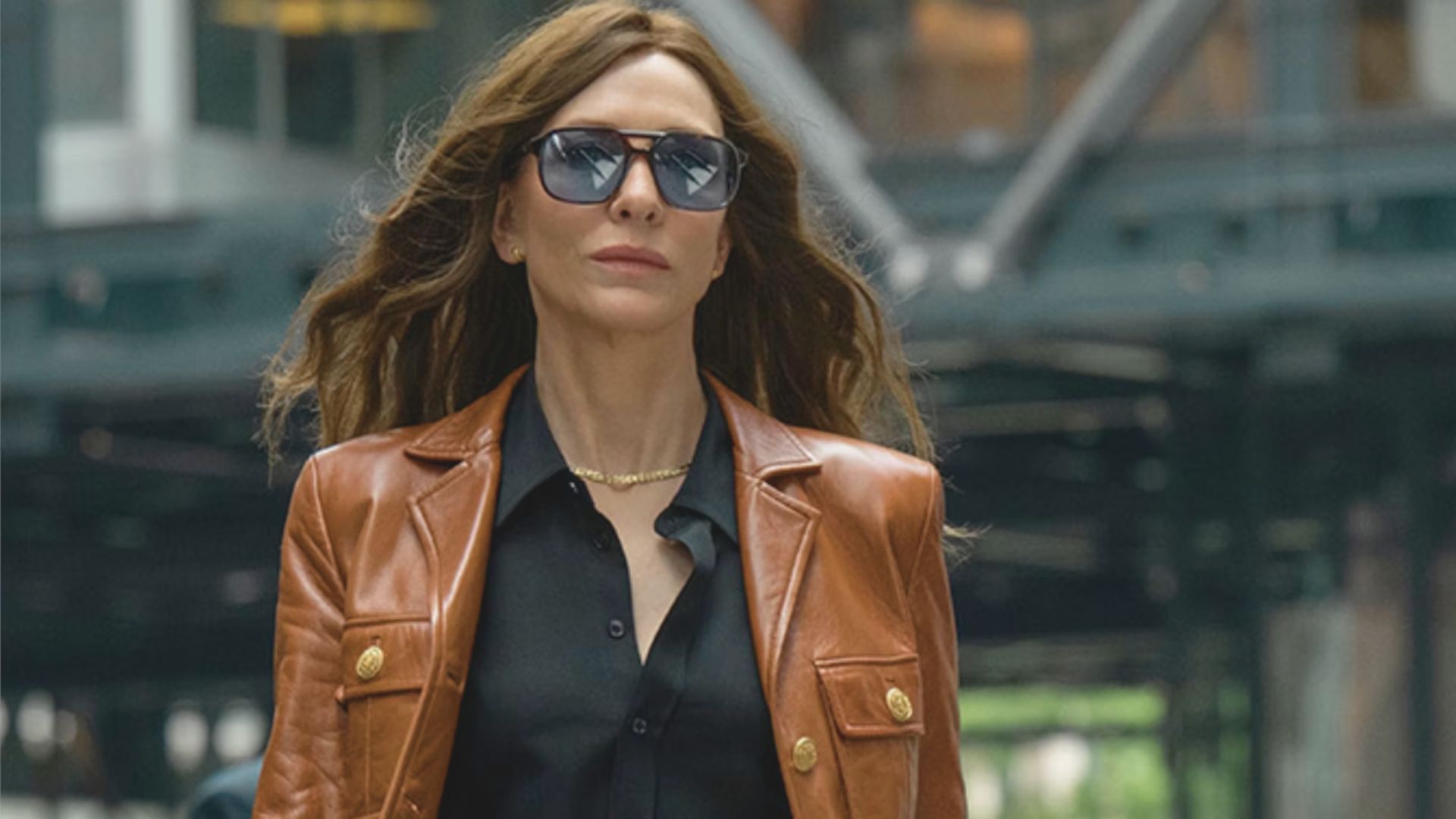 A woman wearing a brown leather jacket and sunglasses stands confidently, exuding a stylish and modern vibe.