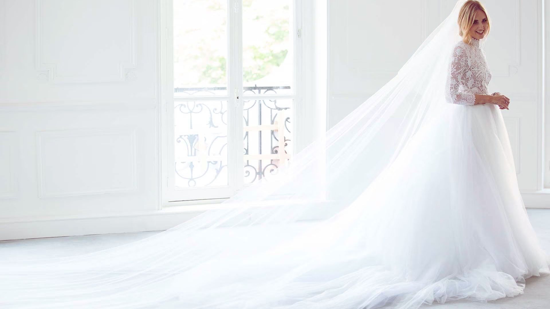 A bride in a long white wedding dress stands gracefully in front of a large window, illuminated by natural light.