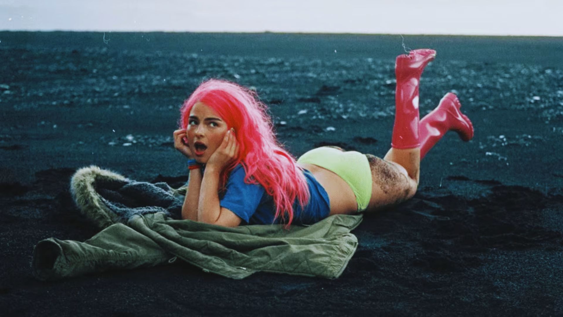 A woman with pink hair relaxing on the beach, lying on a towel under the sun with waves in the background.