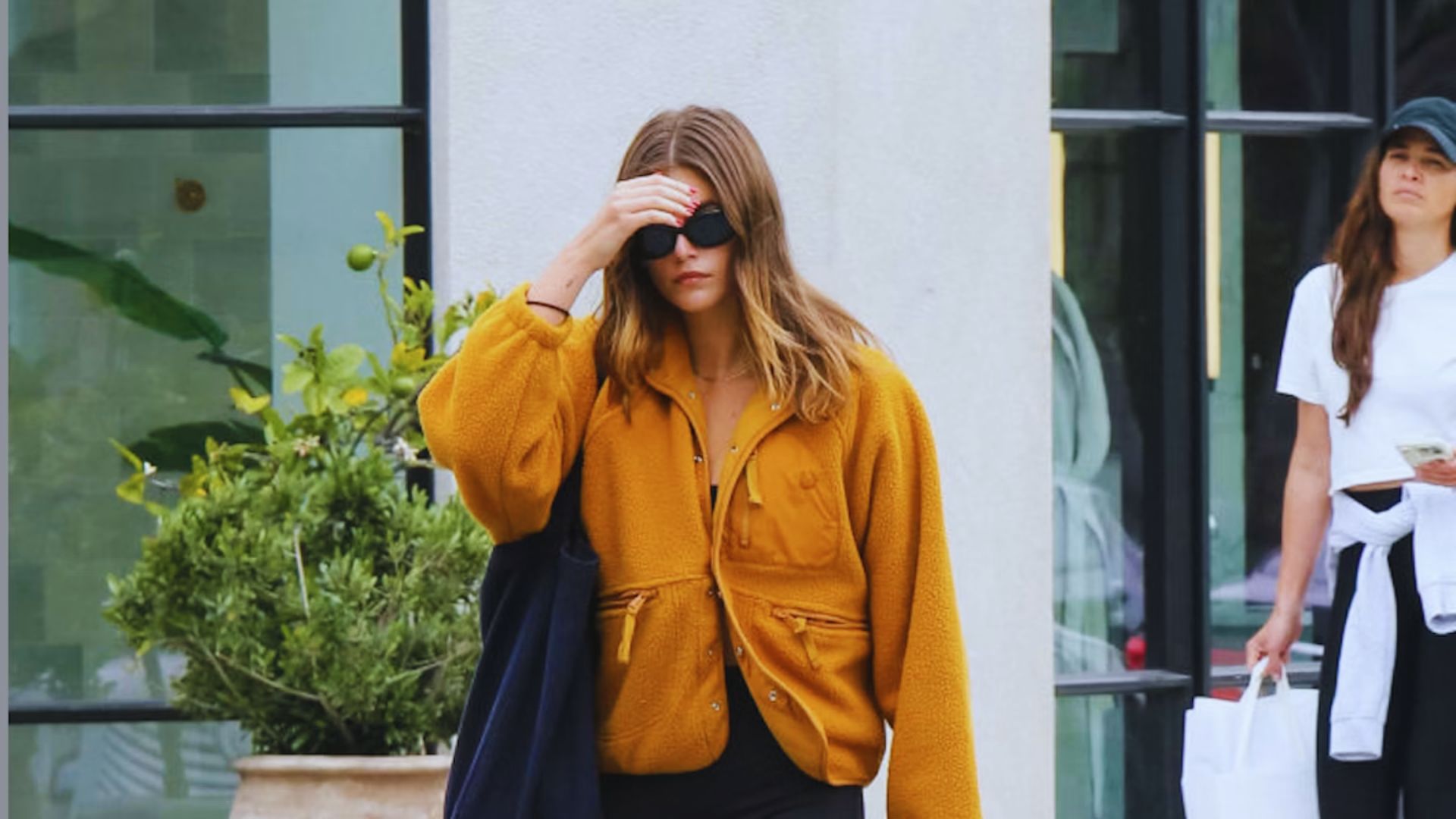 A woman dressed in a mustard jacket and black pants walks down the street, surrounded by urban scenery.