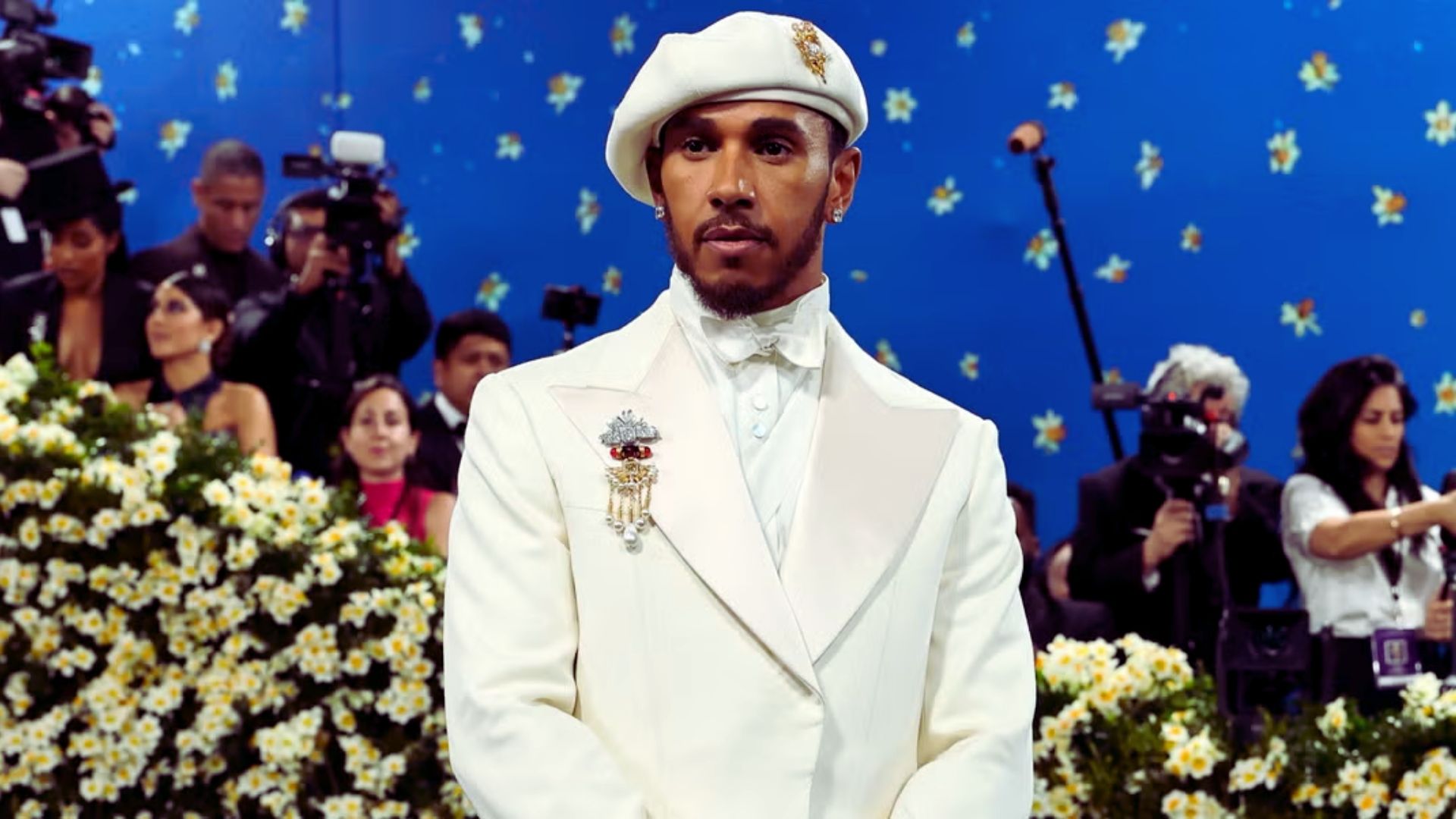 A man wearing a white suit and hat stands amidst a garden of blooming flowers.