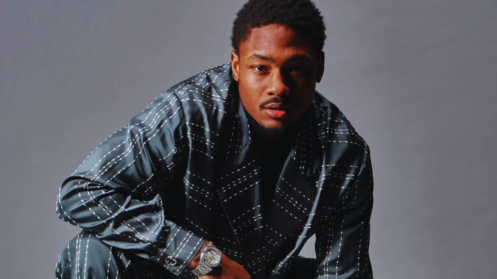 A man in a black and white checkered shirt sits comfortably on a chair, exuding a laid-back demeanor.