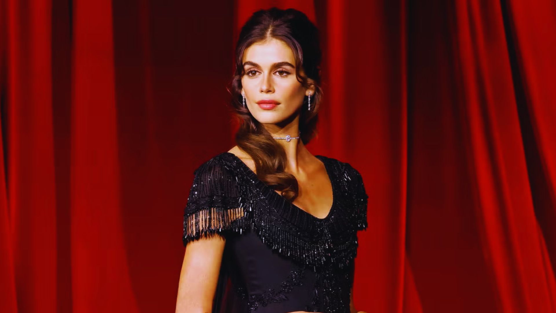 A woman wearing a black dress poses in front of rich red curtains, creating a striking visual contrast.