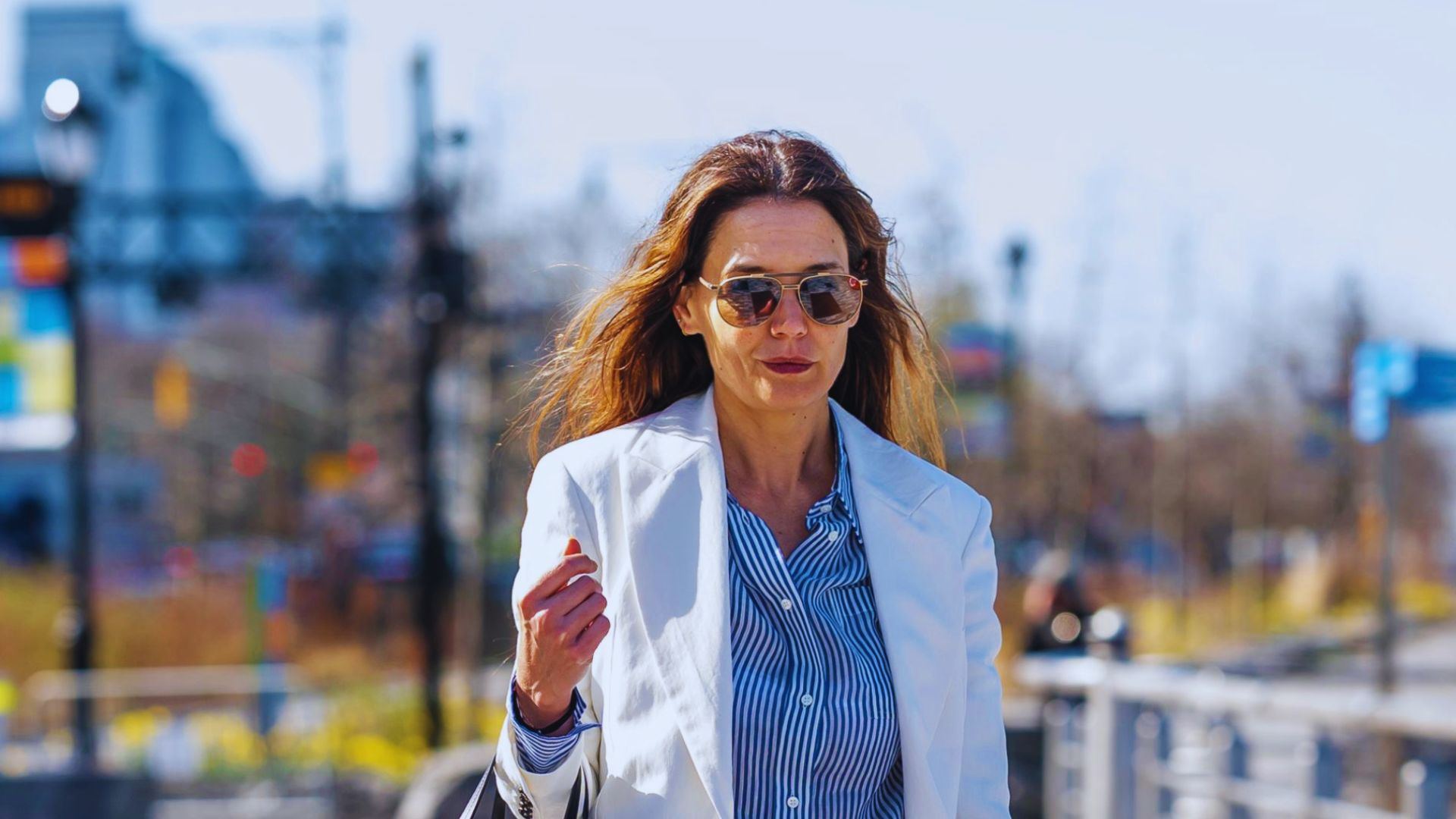 A woman in a white blazer and sunglasses walks down the street, showcasing a chic and modern fashion sense.