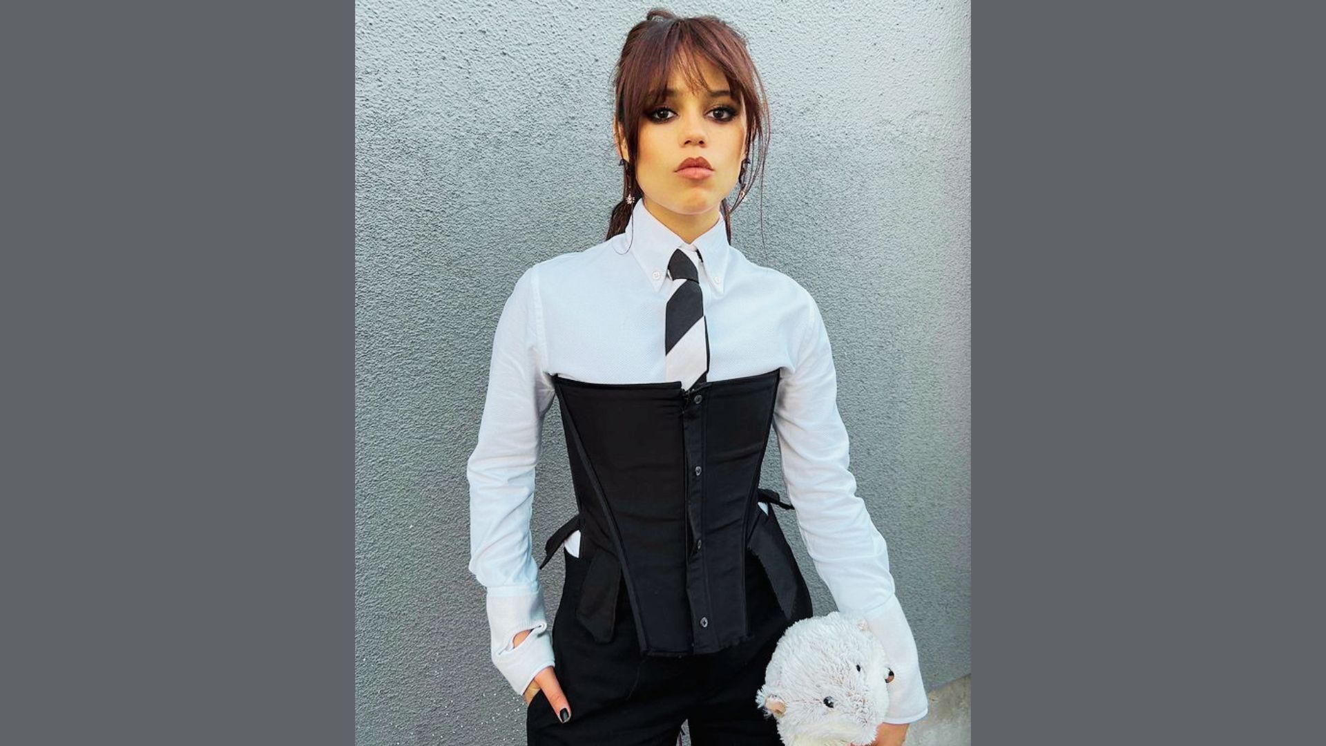 A woman wearing a corset over a t-shirt and a tie, standing confidently against a wall.