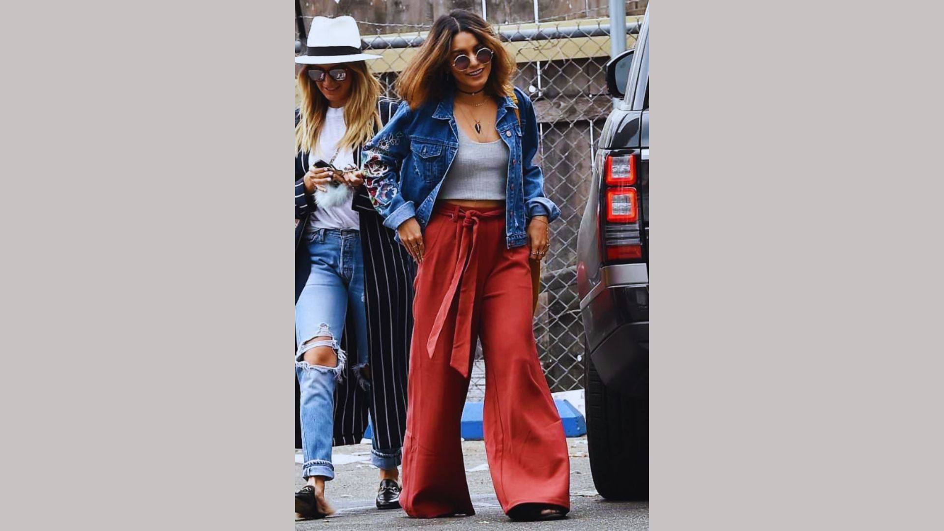 Two women dressed in red pants and denim jackets stroll together along a bustling street.