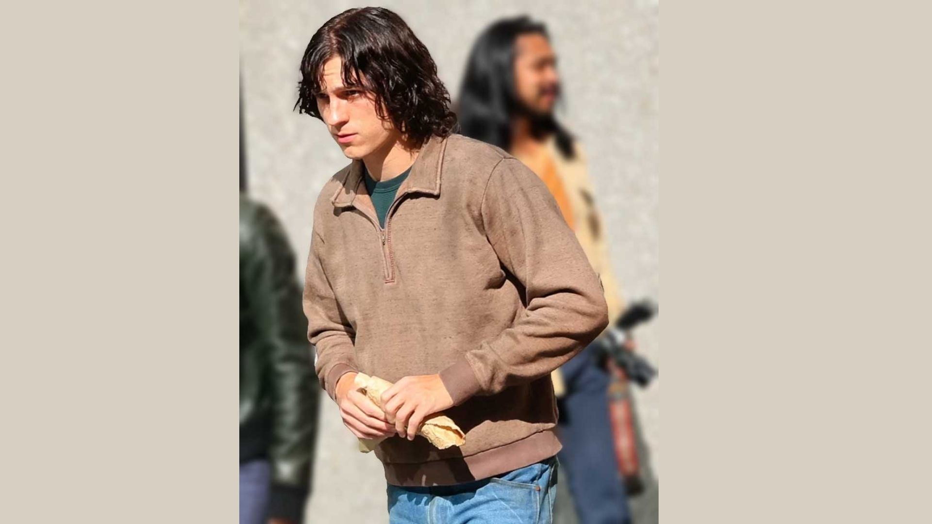 A young man with long hair wearing a brown jacket, hoodie, and slim jeans in a casual off-duty outfit.