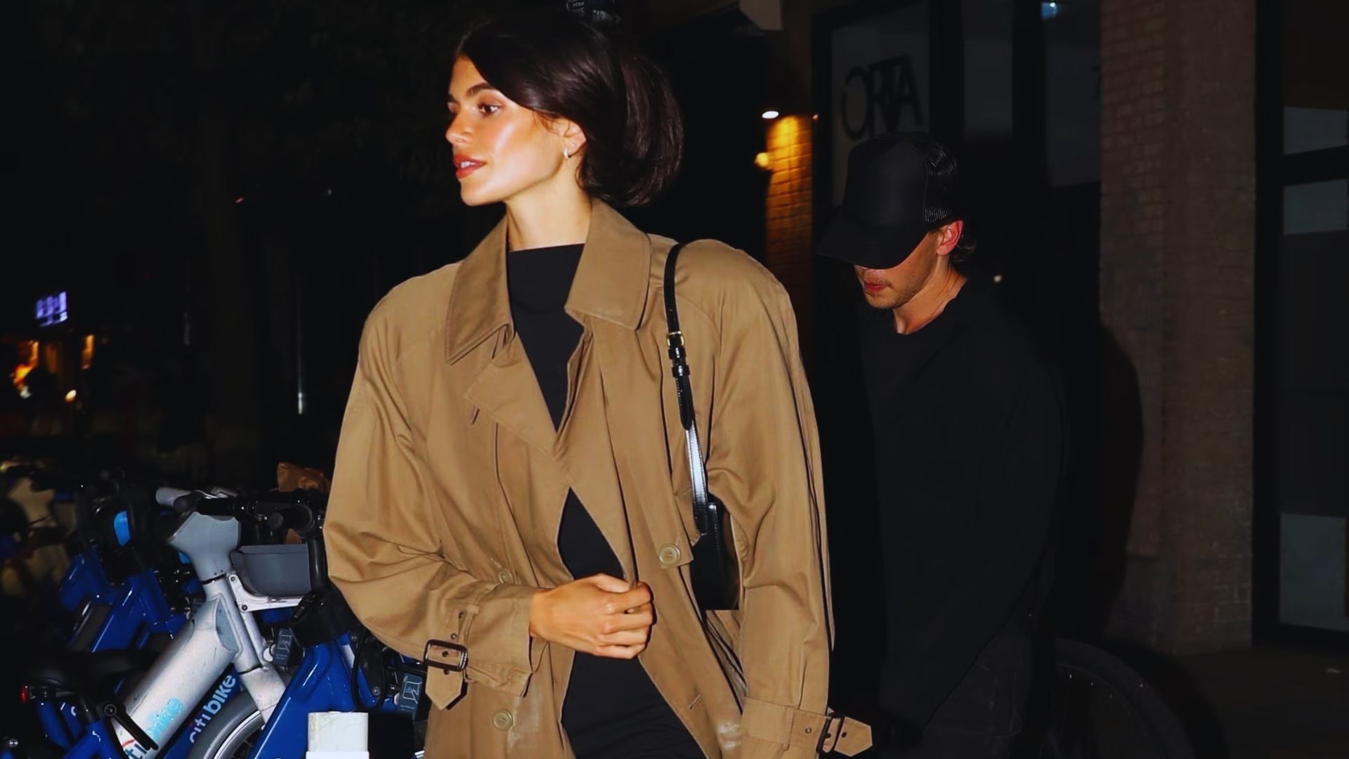 A woman wearing a trench coat strolls along a city street, with shops and pedestrians visible in the background.