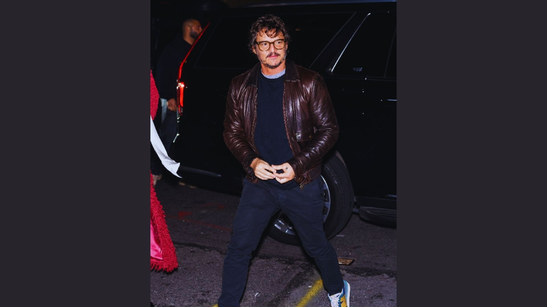 A man in a brown leather jacket and blue jeans stands, embodying a casual style with a touch of ruggedness.