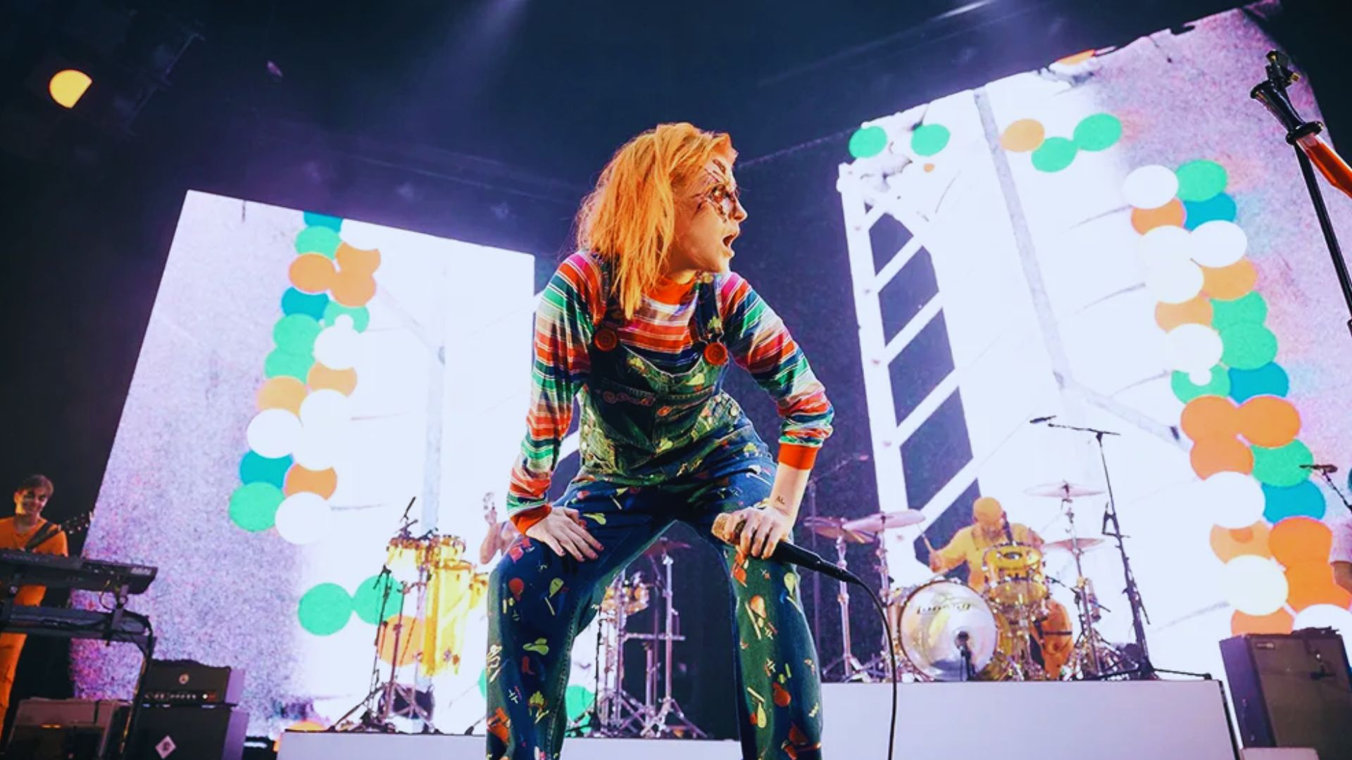 A woman with vibrant orange hair stands on stage, holding a microphone and engaging with the audience.