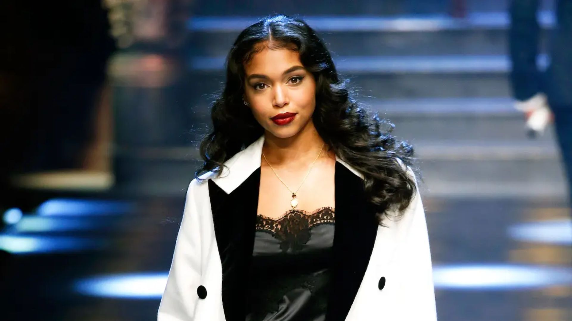 A woman in a black and white dress and jacket confidently walks down a fashion runway.