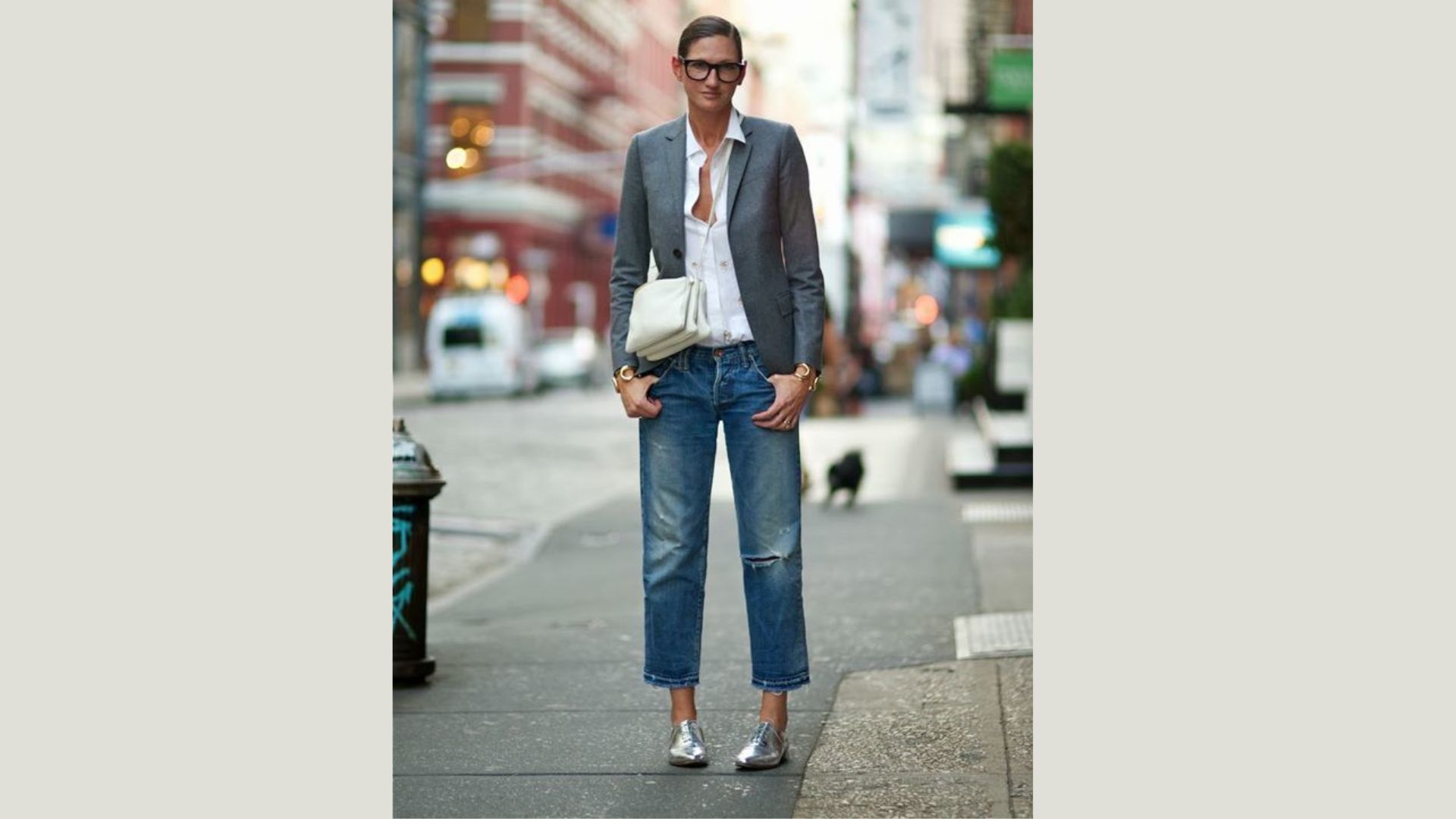 A woman in jeans and a blazer stands confidently on a bustling city street, surrounded by urban architecture.