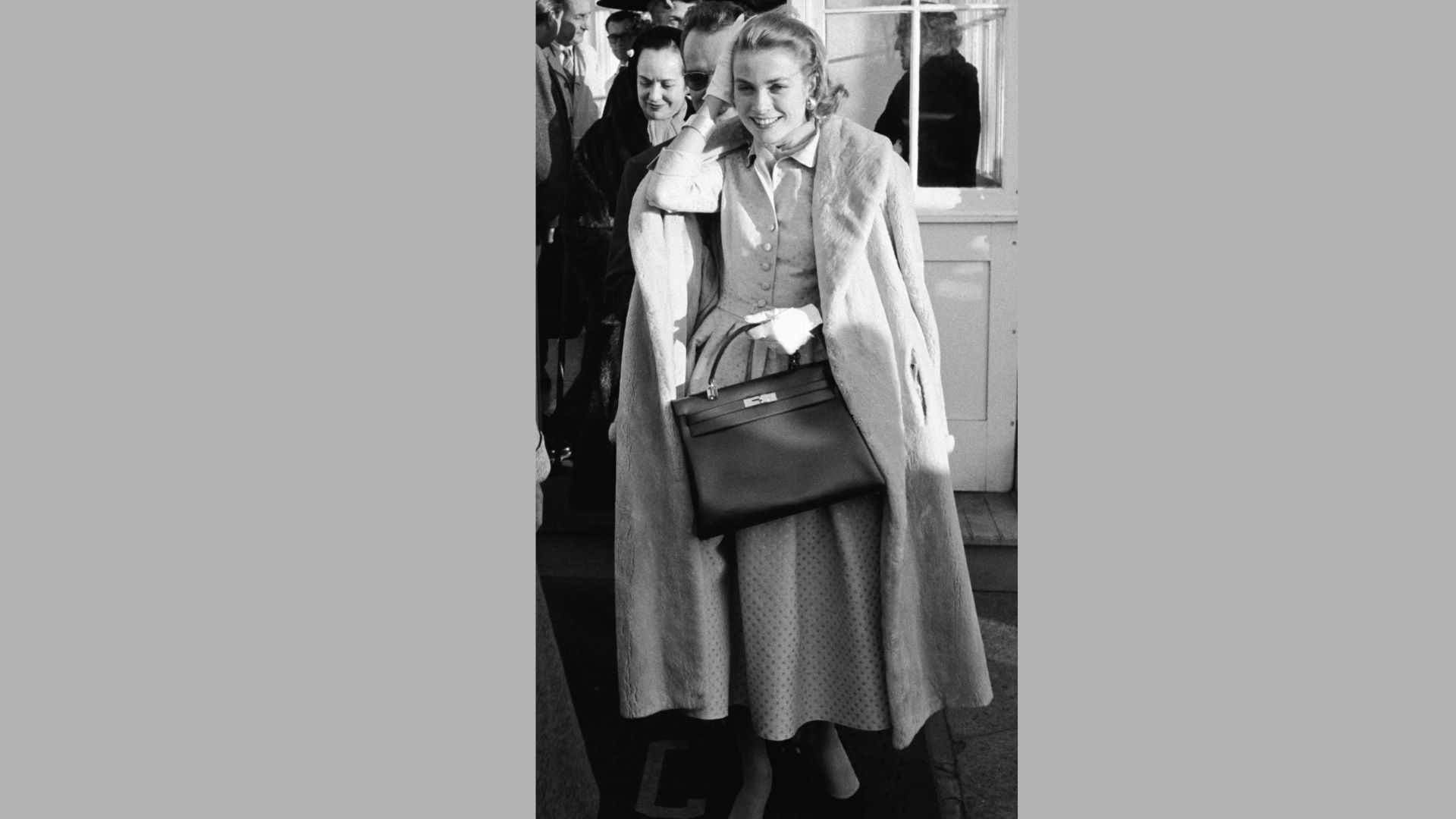 1. A woman wearing a coat and dress holds a purse, standing confidently with a stylish appearance.