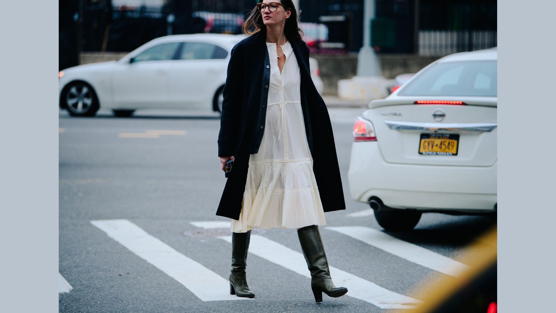 A woman in a white dress and boots strides confidently across the street, embodying a chic urban style.