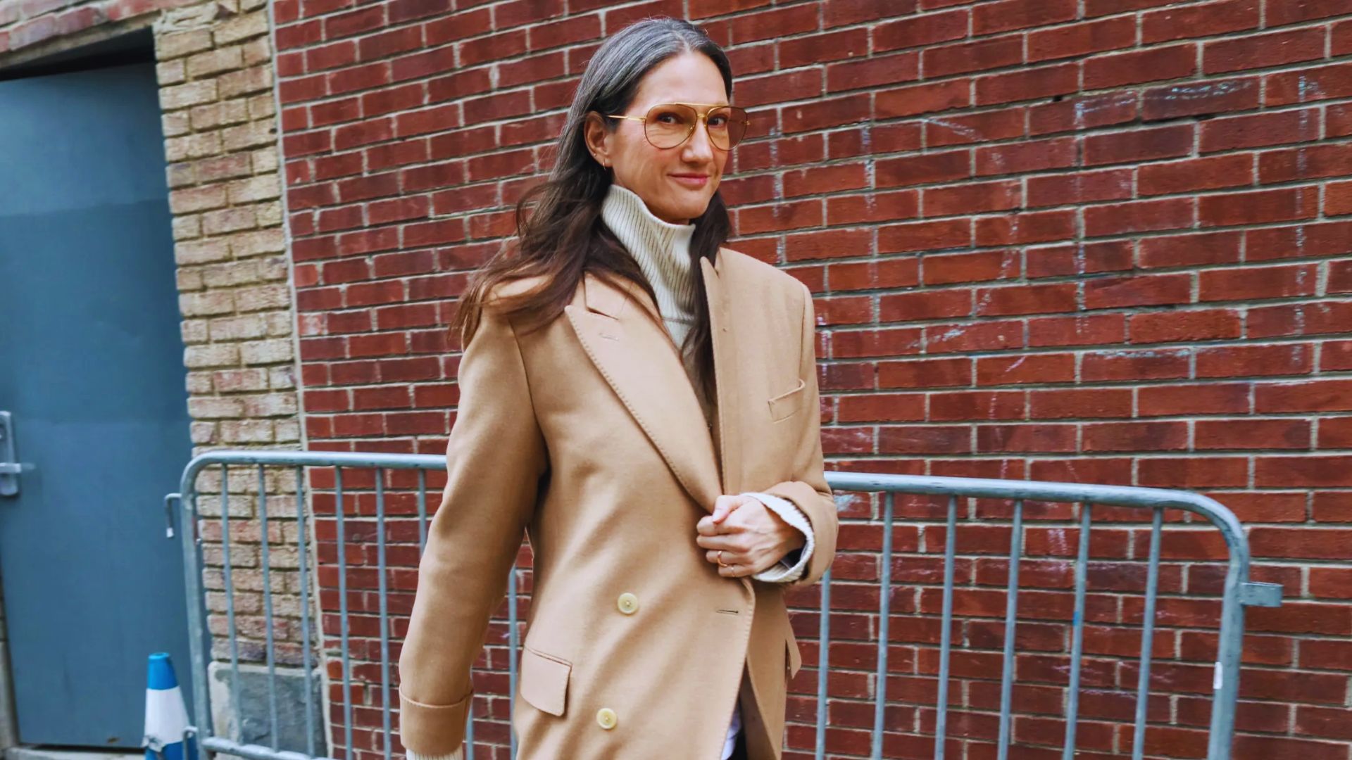 A woman with glasses dons a camel coat, exuding sophistication and style in her appearance.