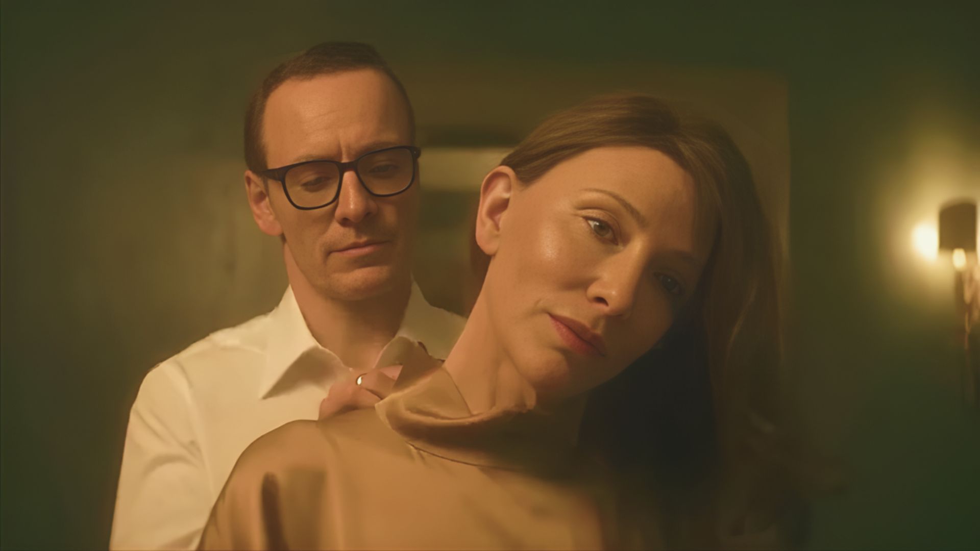 A man and woman converse in a room, while a man in glasses observes them from a distance.