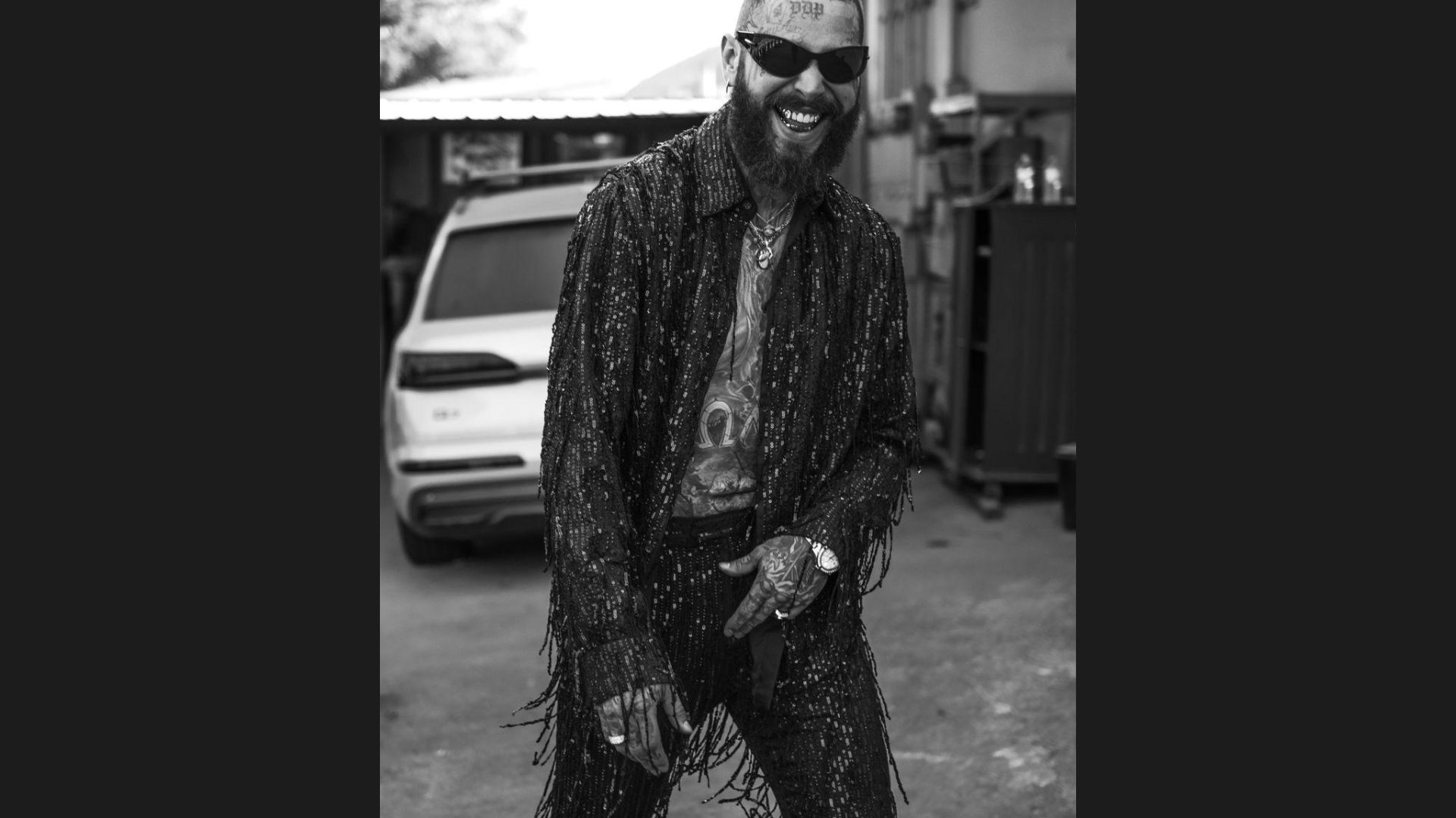 A bearded man wearing sunglasses stands confidently in front of a parked car.