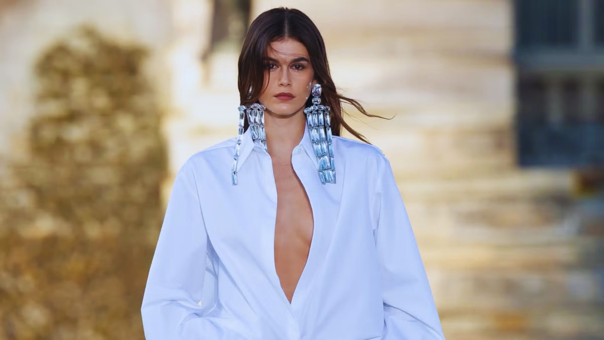 A model walks the runway in a white shirt paired with striking blue earrings, showcasing a chic fashion look.