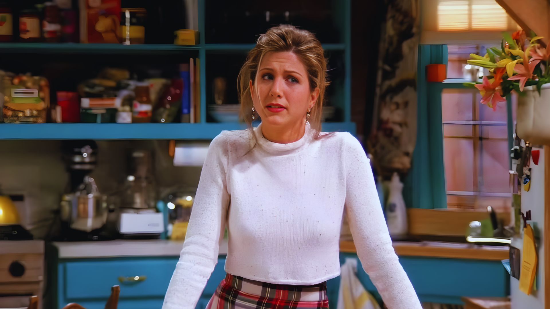 A woman wearing a plaid skirt stands in a modern kitchen, surrounded by cooking utensils and appliances.