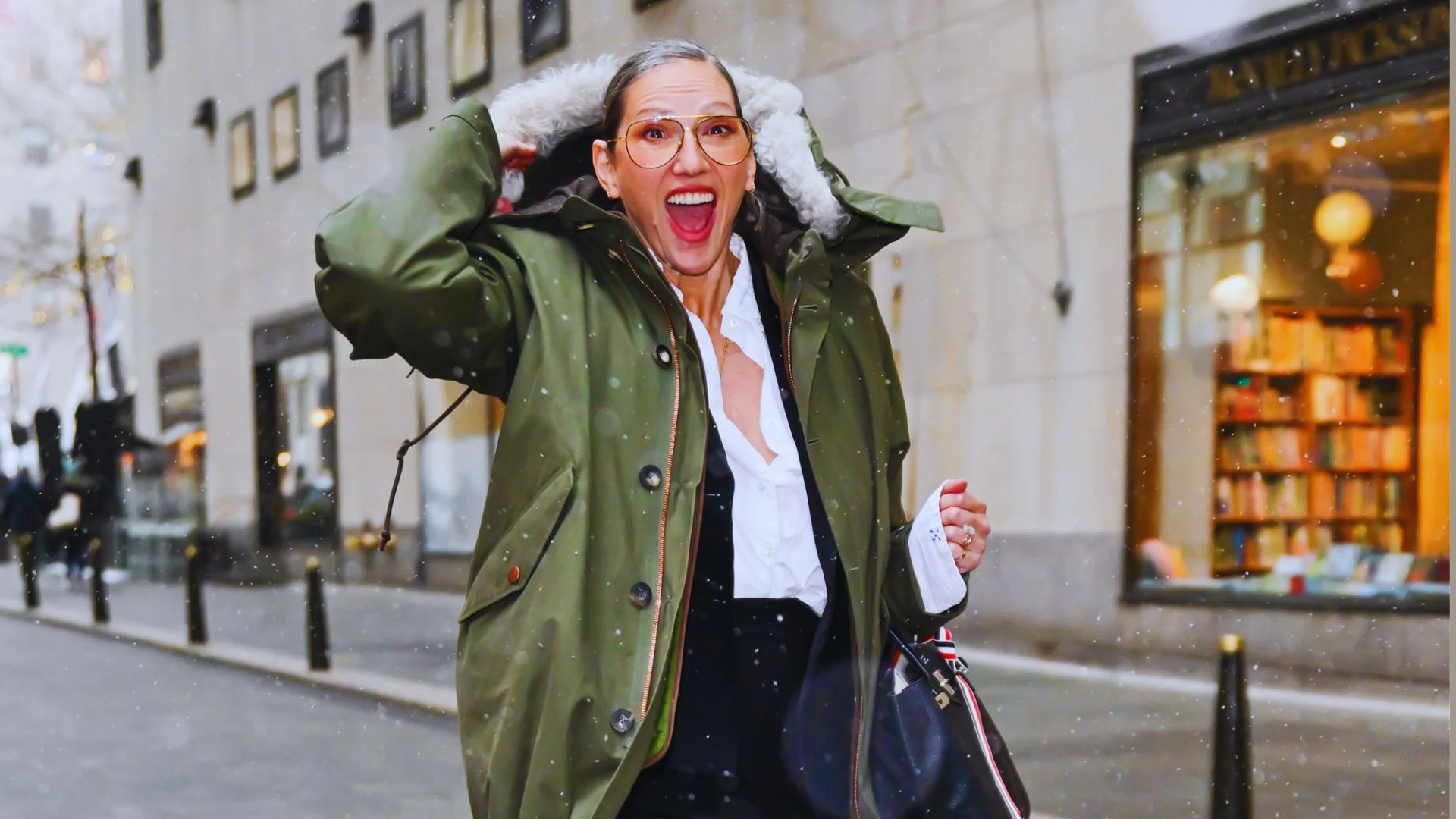 A woman wearing a green coat and black pants laughs heartily, showcasing her joyful expression.