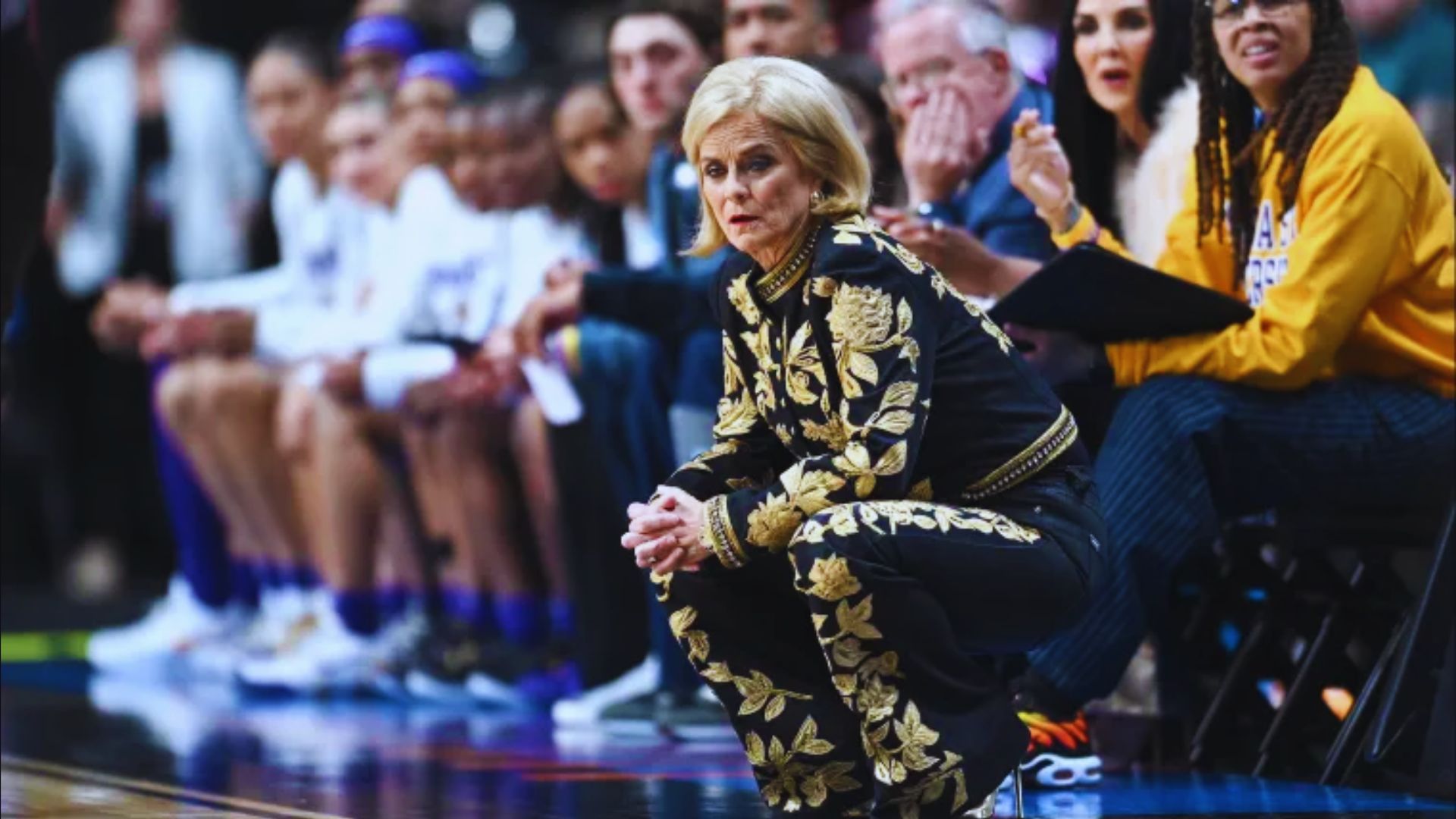 A woman sitting on the sidelines, hands resting on her knees, appears contemplative and engaged in the moment.