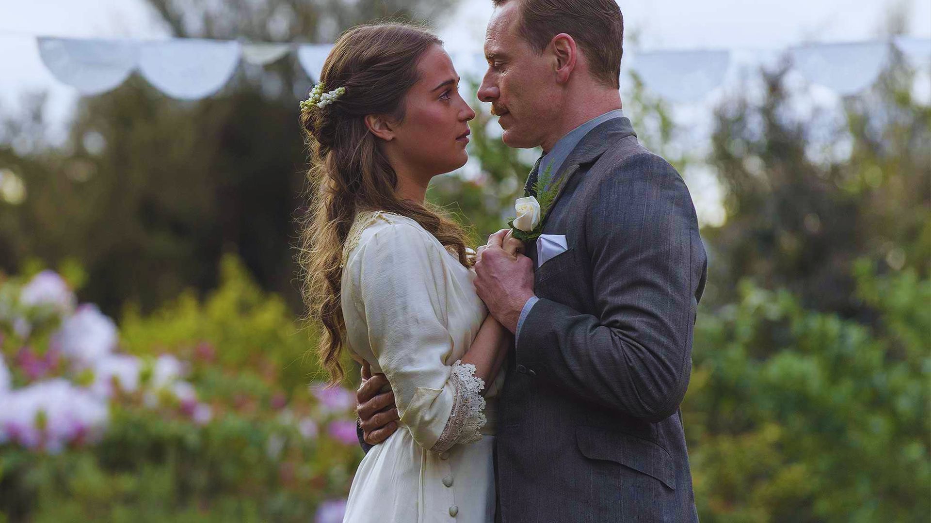 A man and woman share a kiss in a lush garden, surrounded by vibrant flowers and greenery.