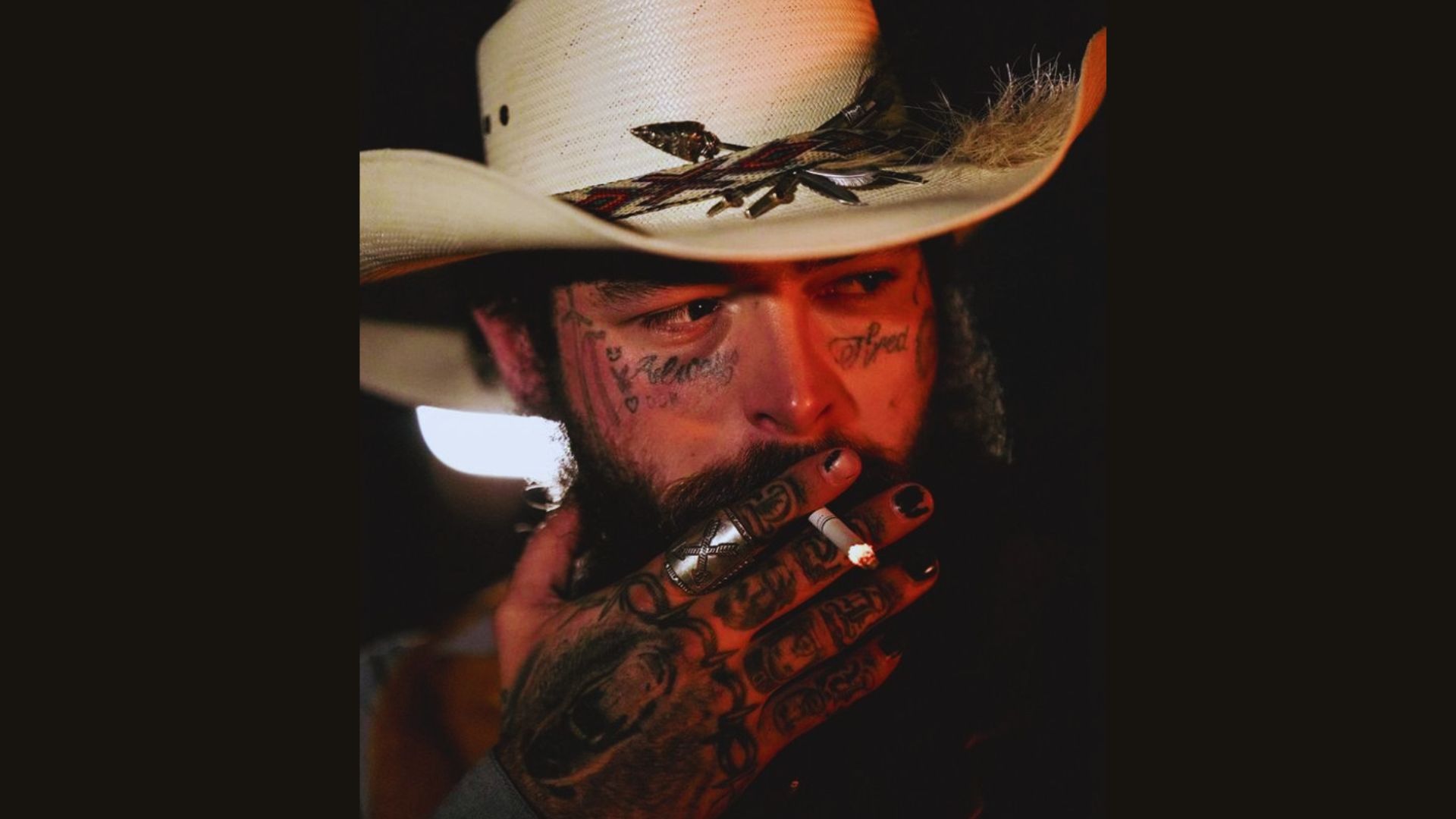 A man with tattoos wearing a cowboy hat, smoking a cigarette, stands confidently against a blurred background.