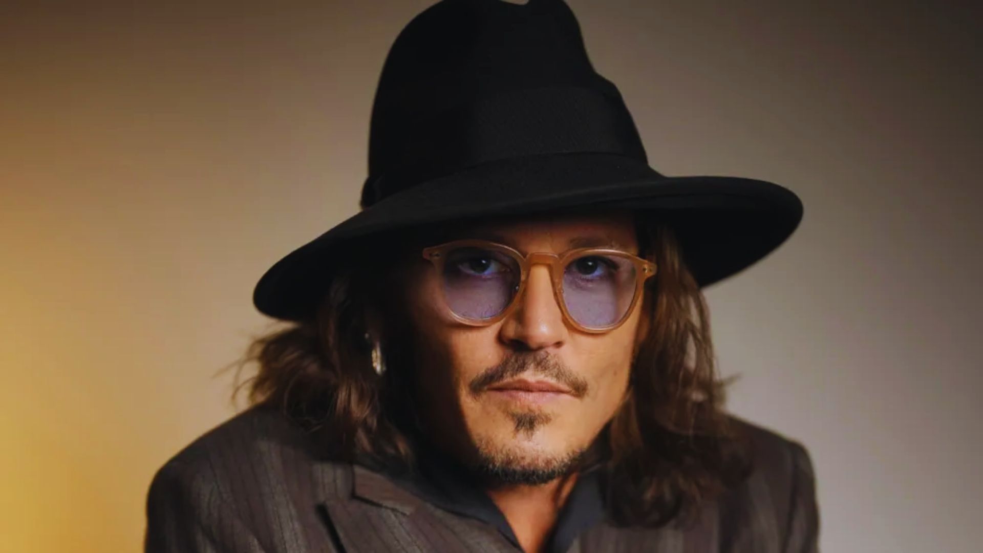 Johnny Depp wearing a stylish hat and glasses, looking thoughtfully off to the side.