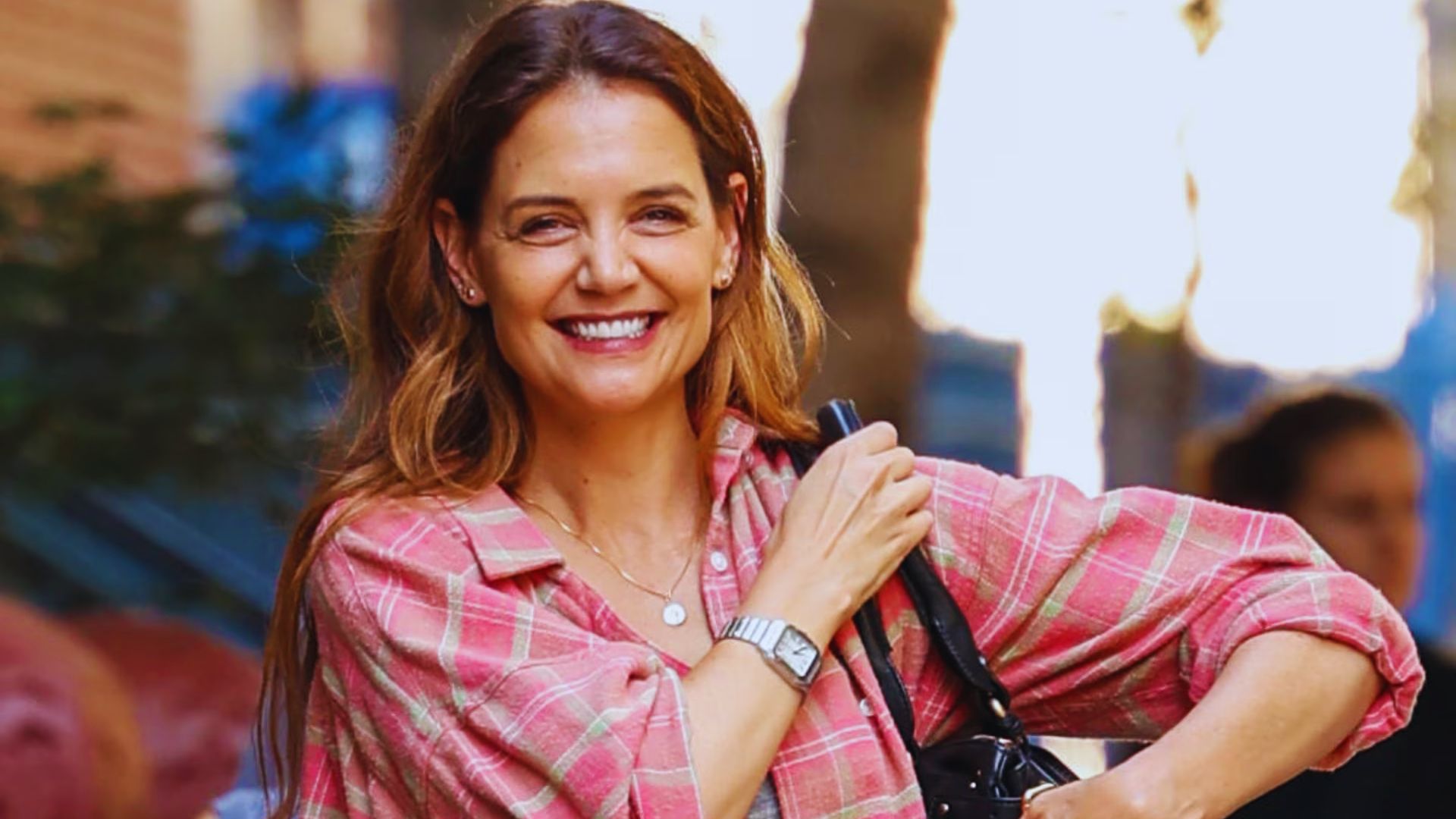 A smiling woman in a pink shirt holds a bag, exuding a cheerful and friendly demeanor.