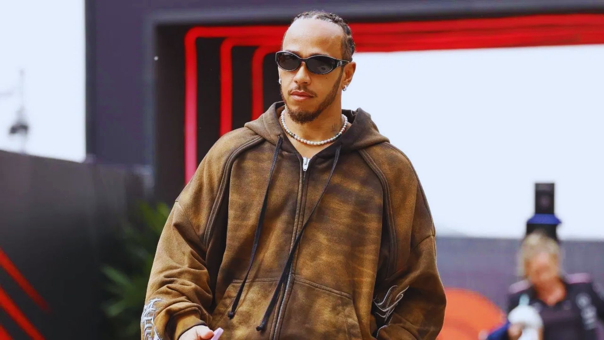 A man wearing a brown hoodie and sunglasses stands confidently, showcasing a casual and stylish look.