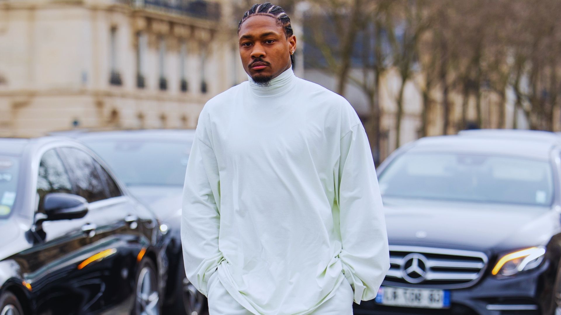 A man dressed in white clothing stands on a city street, surrounded by urban scenery.