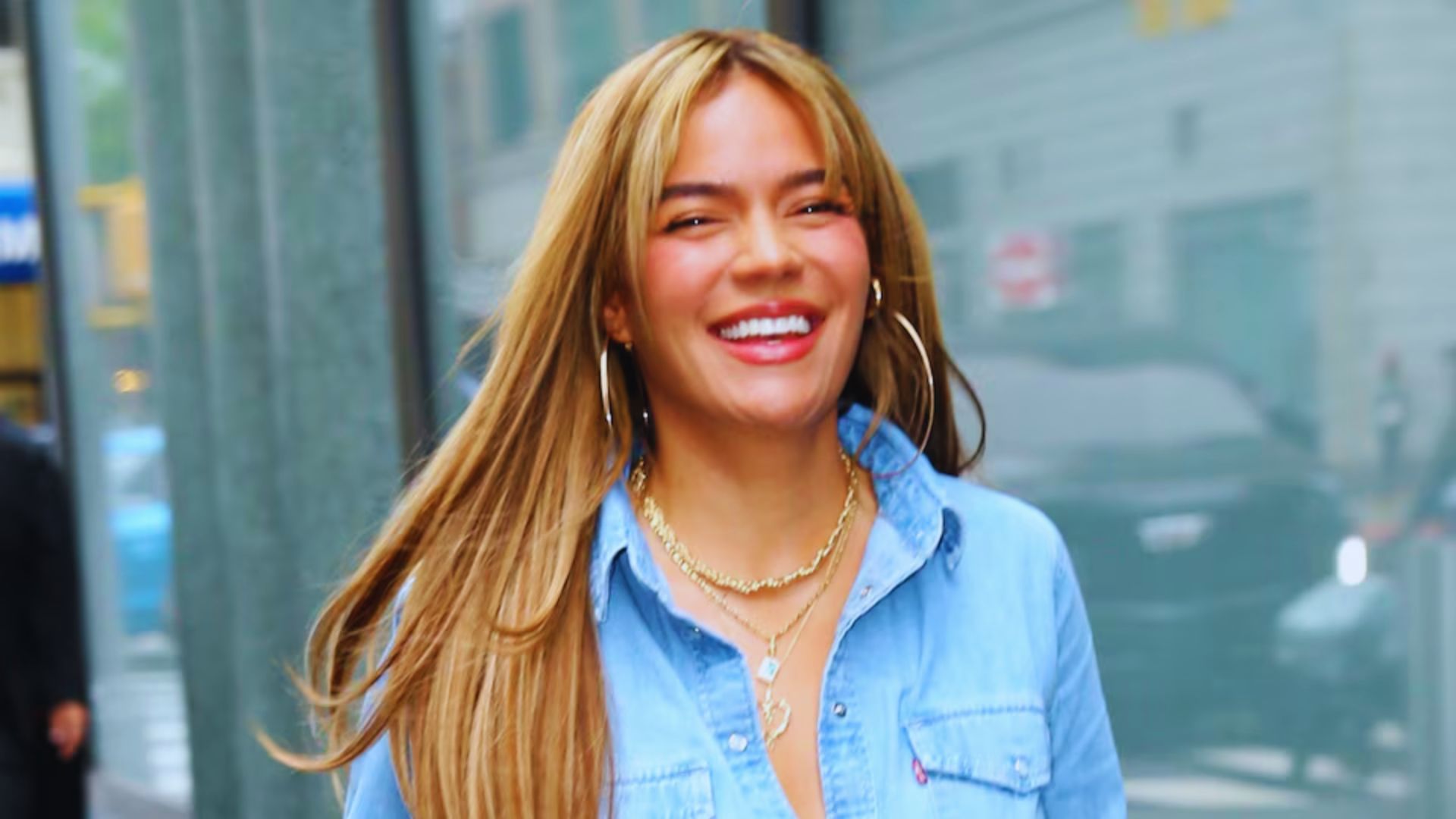 A woman with long hair wearing a denim shirt, standing against a neutral background.