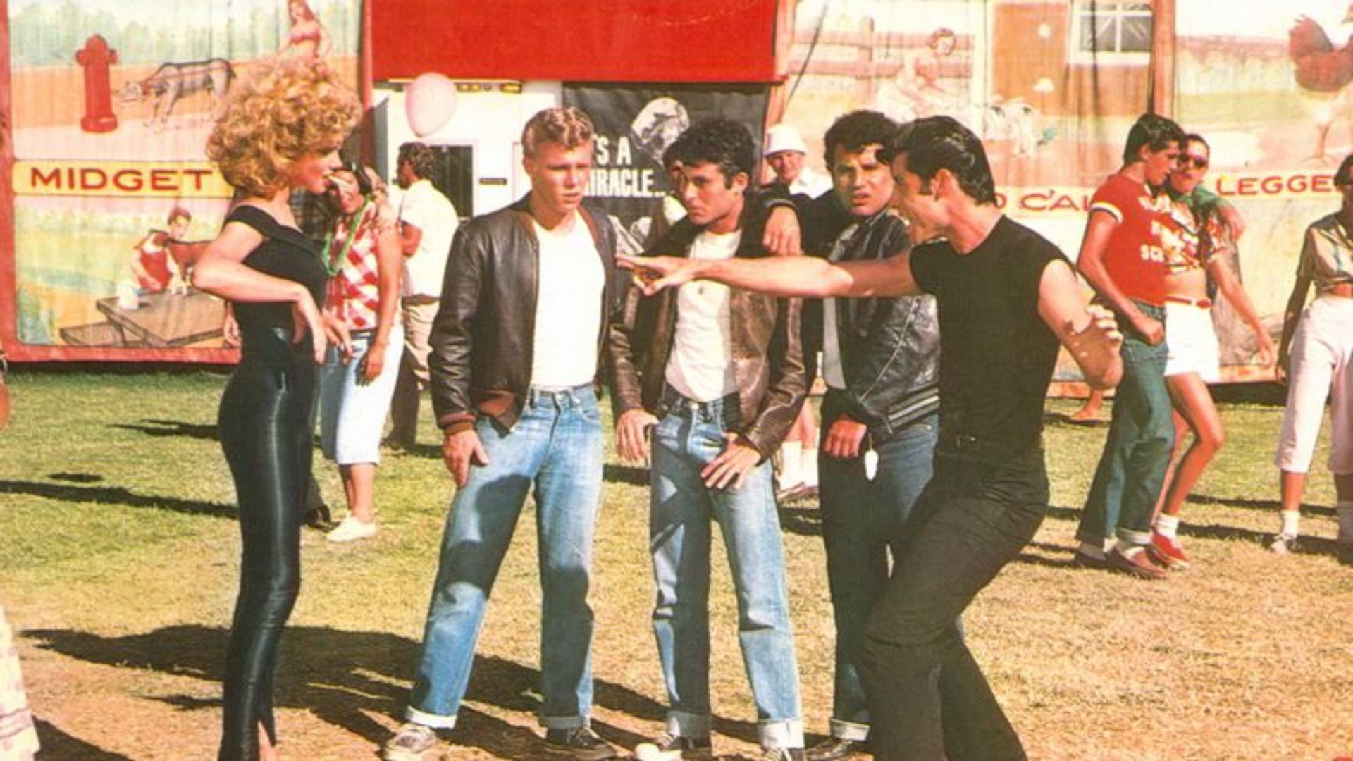 A group of people gathered around a man in a red shirt, dressed in 1950s attire at a themed party from the movie Grease.