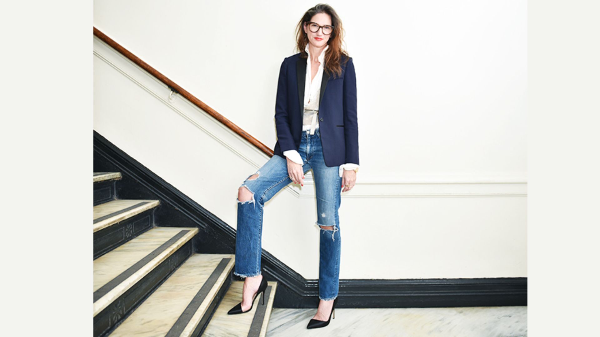 A woman in jeans and a blazer stands confidently on a staircase, showcasing a professional yet casual style.