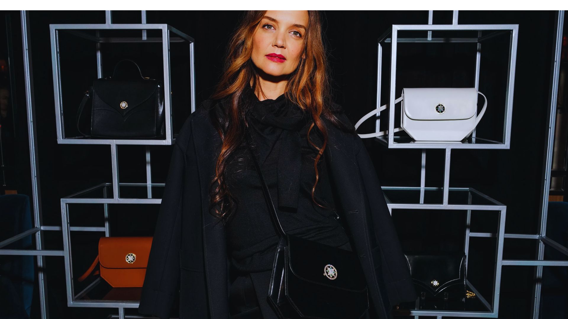 A woman in a black coat and dress stands before a wall displaying various bags.