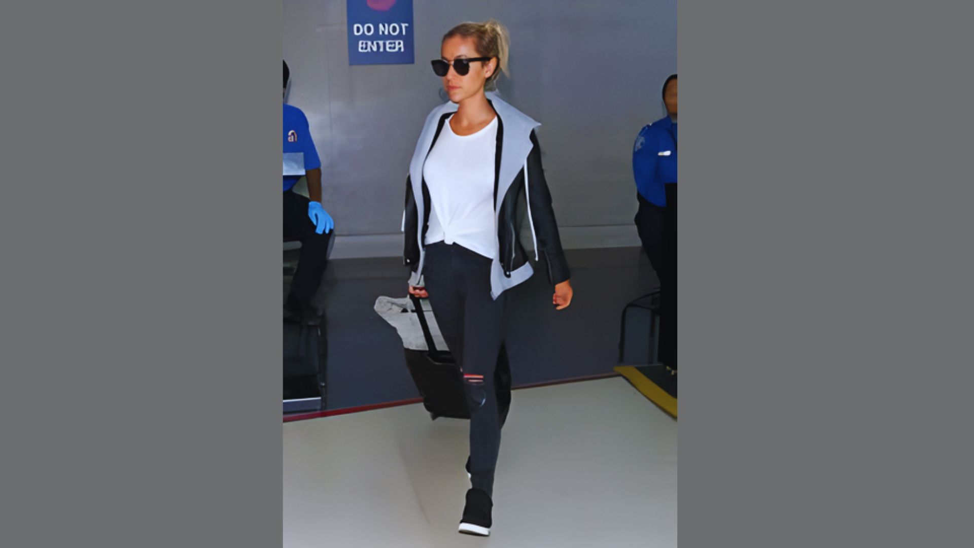 A woman in black jeans and a white shirt walks through an airport terminal, carrying a small bag.