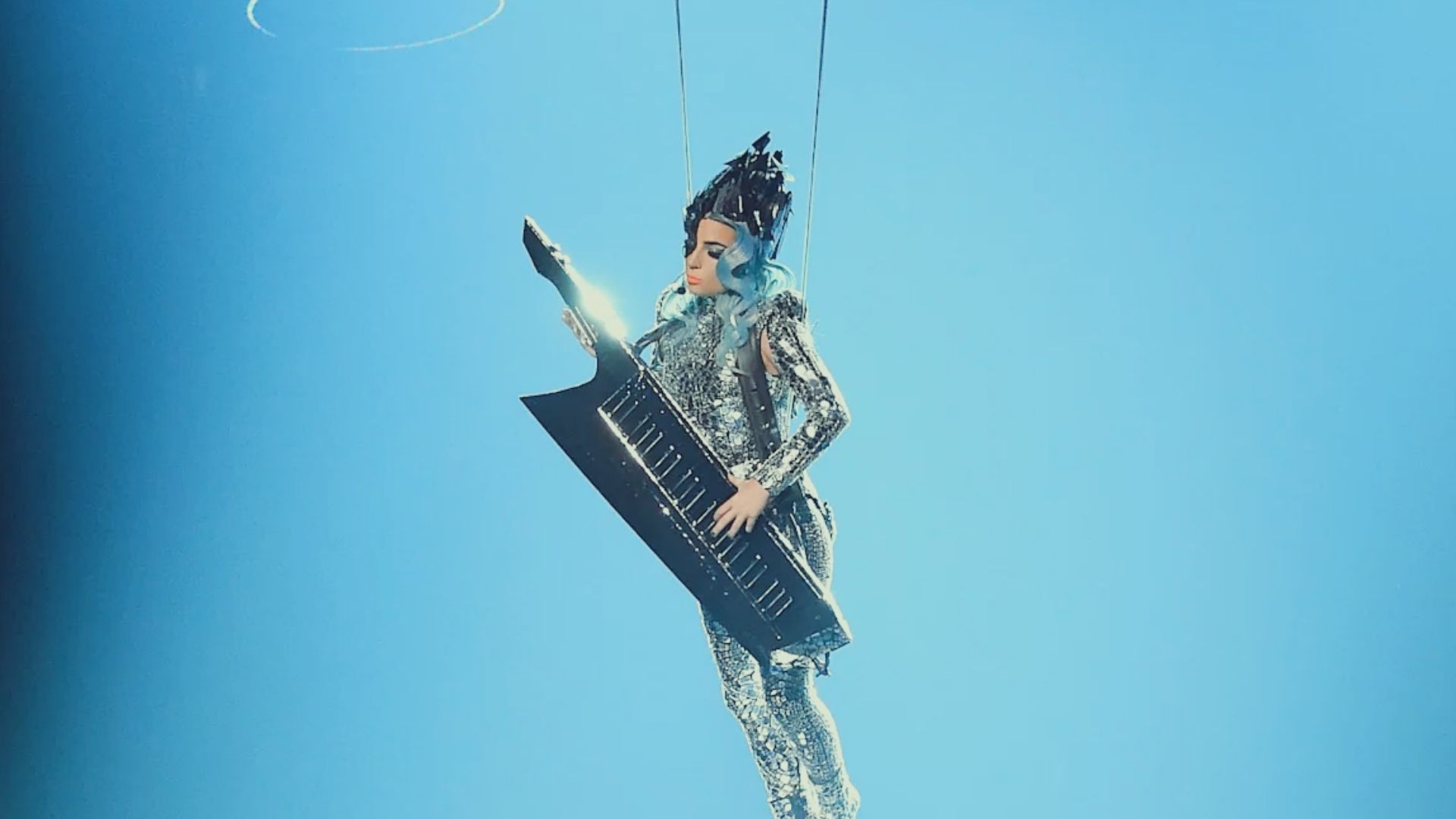 A woman wearing a silver suit hangs from a rope, demonstrating a striking pose in mid-air against a plain backdrop.