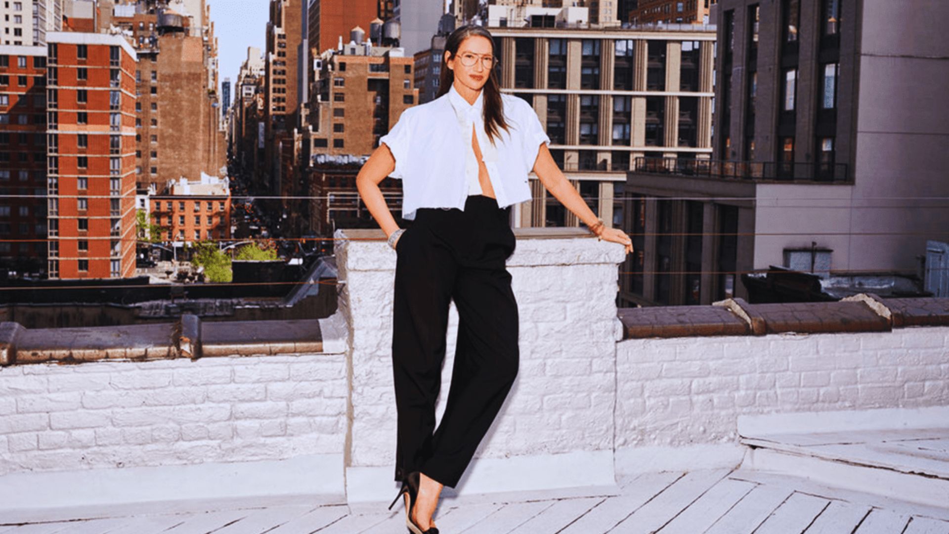 A woman in black pants and a white shirt poses confidently on a rooftop with a city skyline in the background.