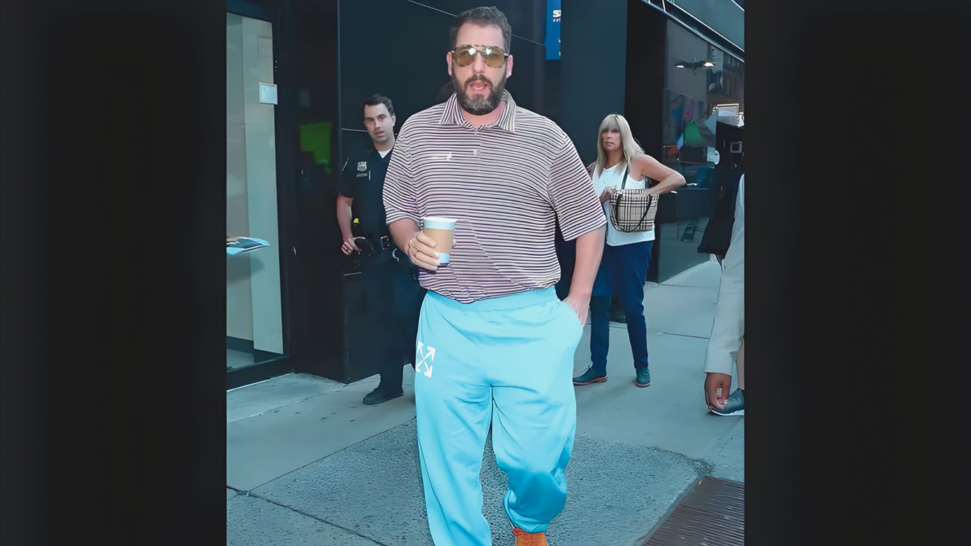 A man in a striped shirt and blue pants walks down the street, showcasing a casual outfit reminiscent of Adam Sandler's style.